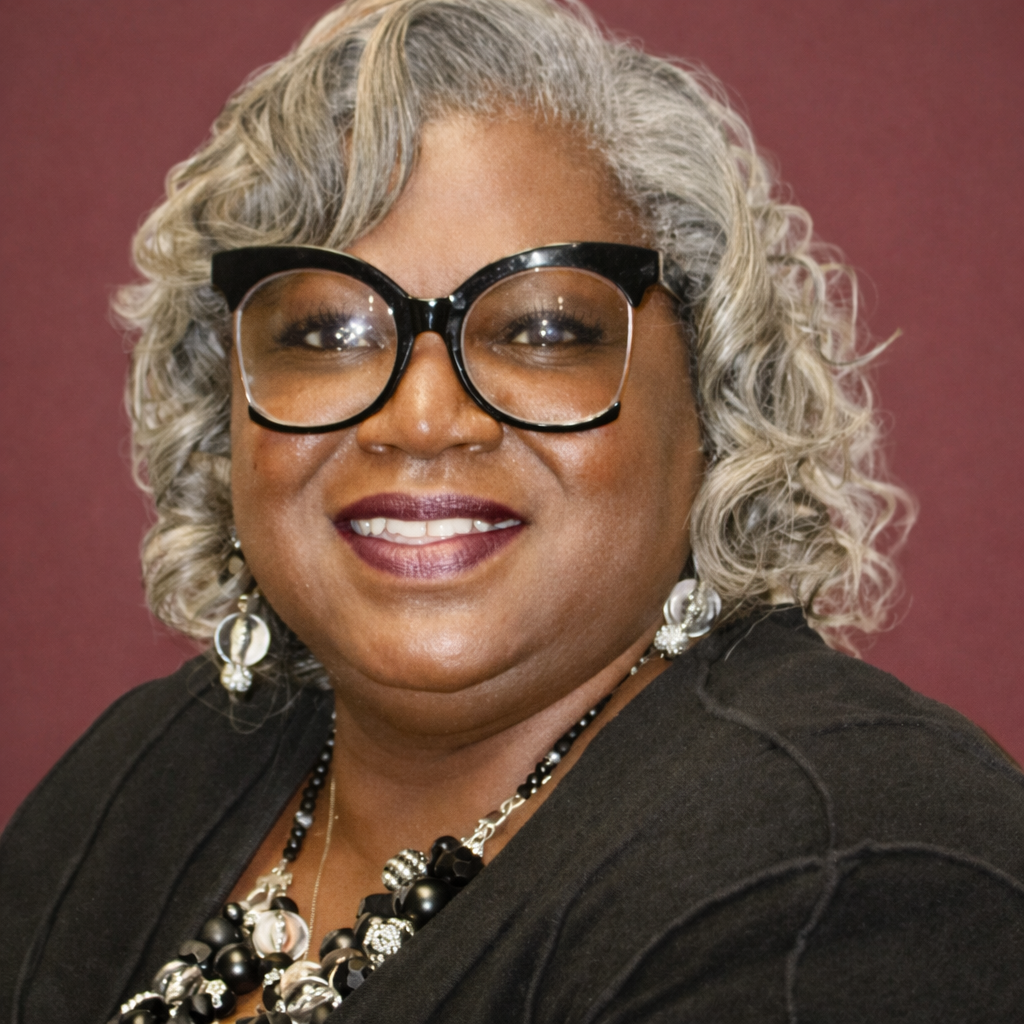 A smiling woman with gray curly hair wearing large black glasses, statement earrings, black and white jewelry, and a black top, against a purple background.