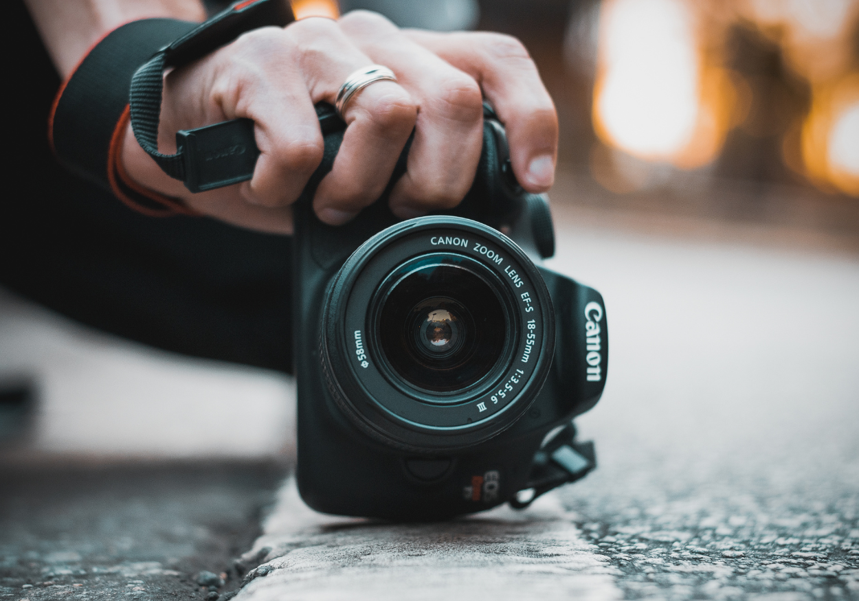 Close-up of a person's hand holding a Canon DSLR camera, with a focus on the camera lens, outdoors with a blurred warm-toned background.