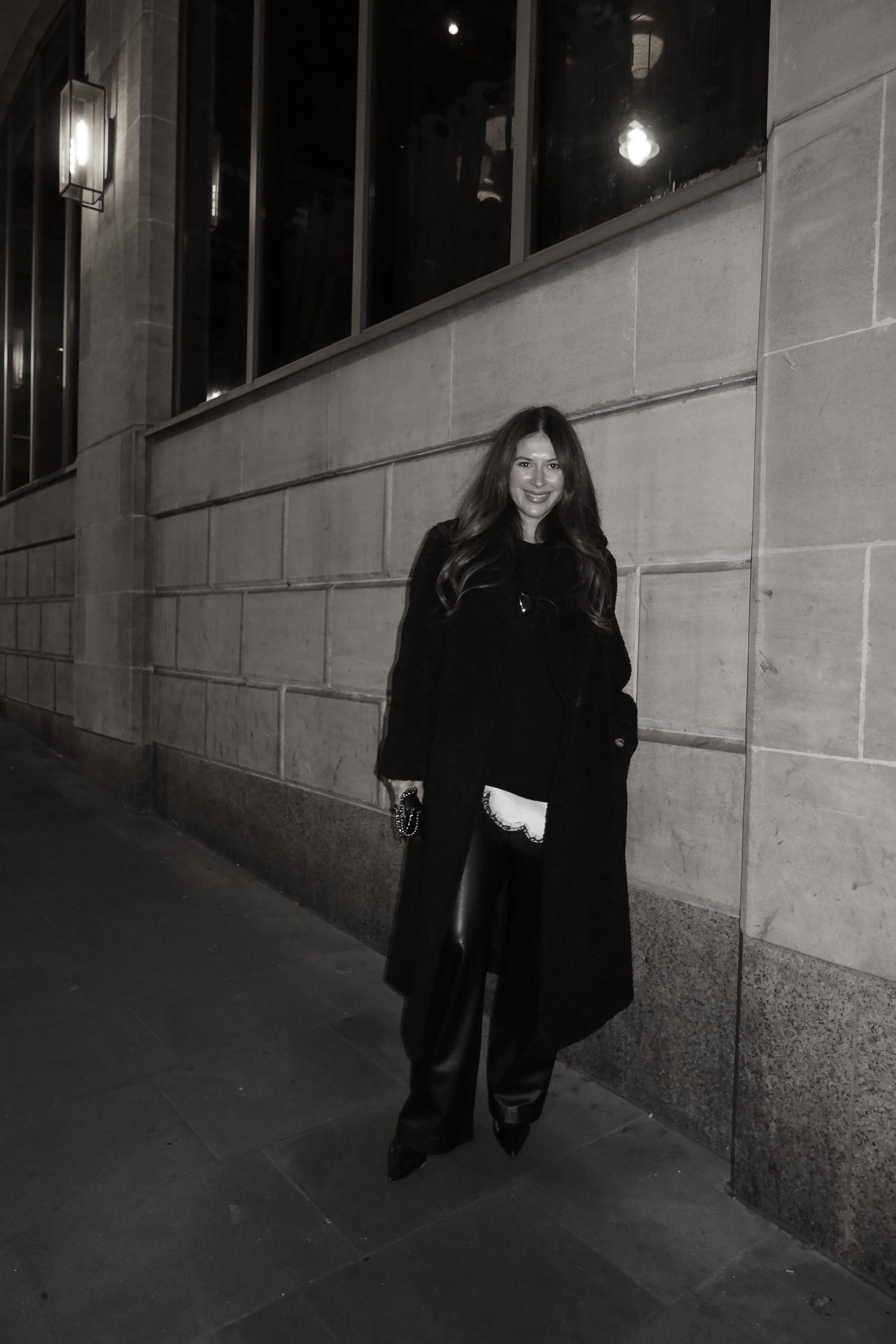 A woman with long, wavy hair smiling and standing on a city sidewalk at night, wearing a black coat and shiny black pants, with illuminated streetlights and windows in the background.