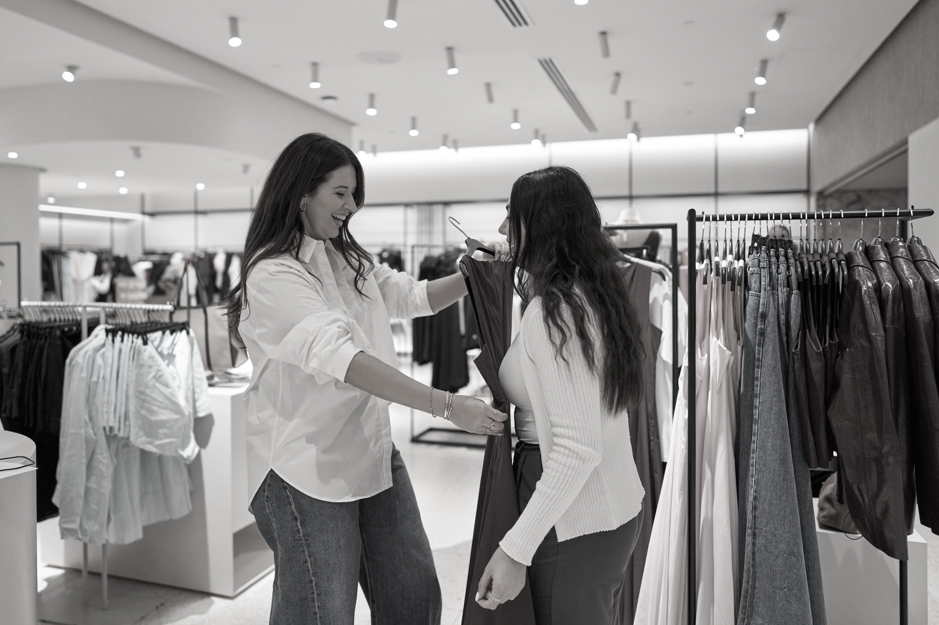 Two women shopping and trying clothes in a store, smiling and engaging with each other.