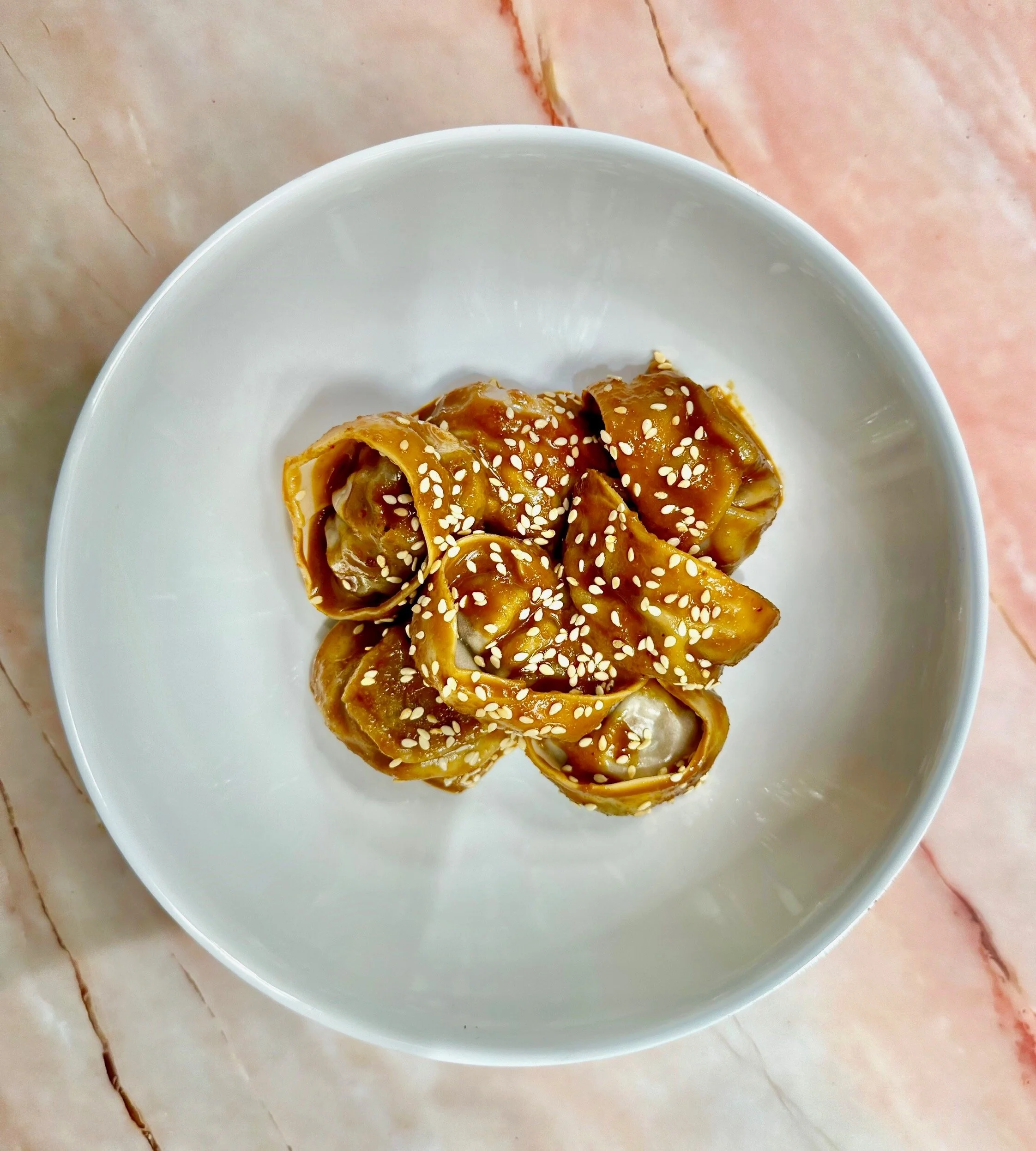 Close-up of six cooked dumplings topped with sesame seeds in a white bowl on a marble surface.