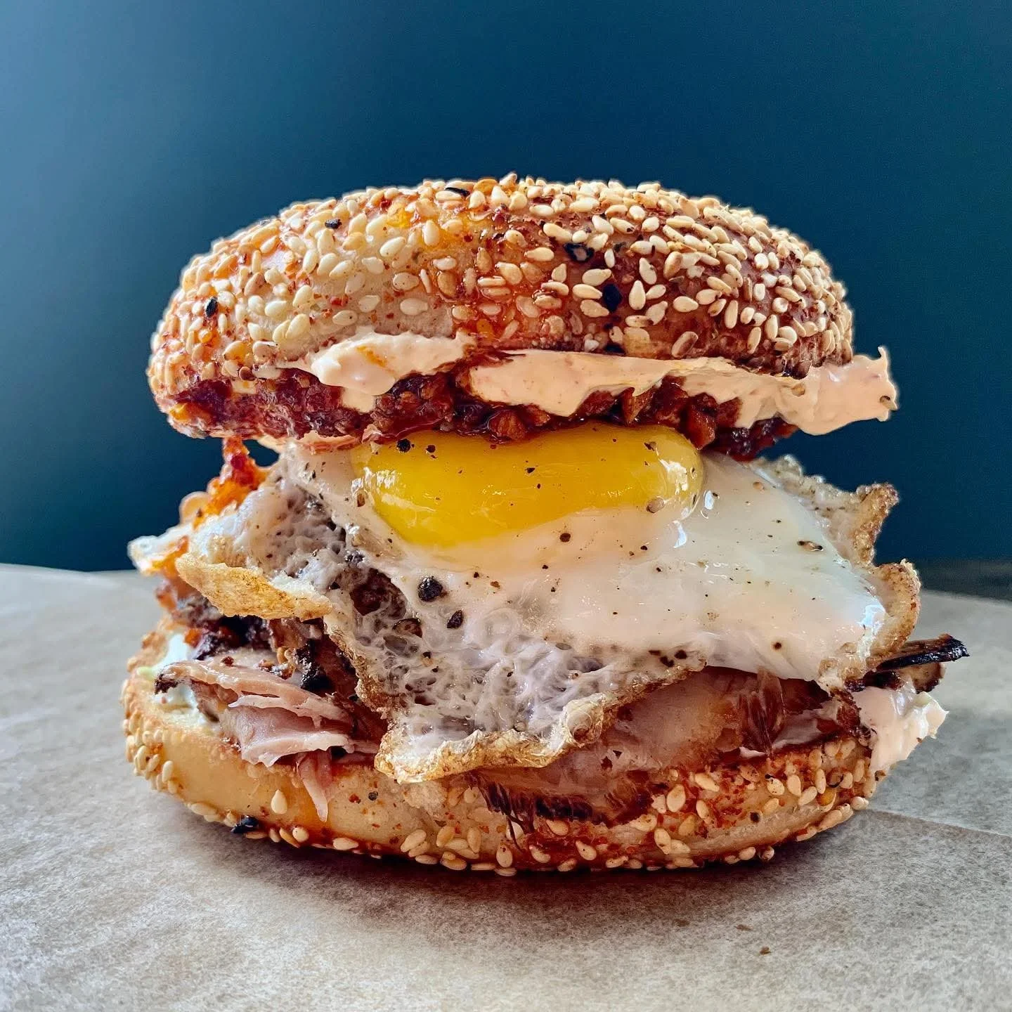 Close-up of a breakfast sandwich with a bagel bun, fried egg, slice of ham, a fried egg, and cream cheese, topped with sesame seeds.