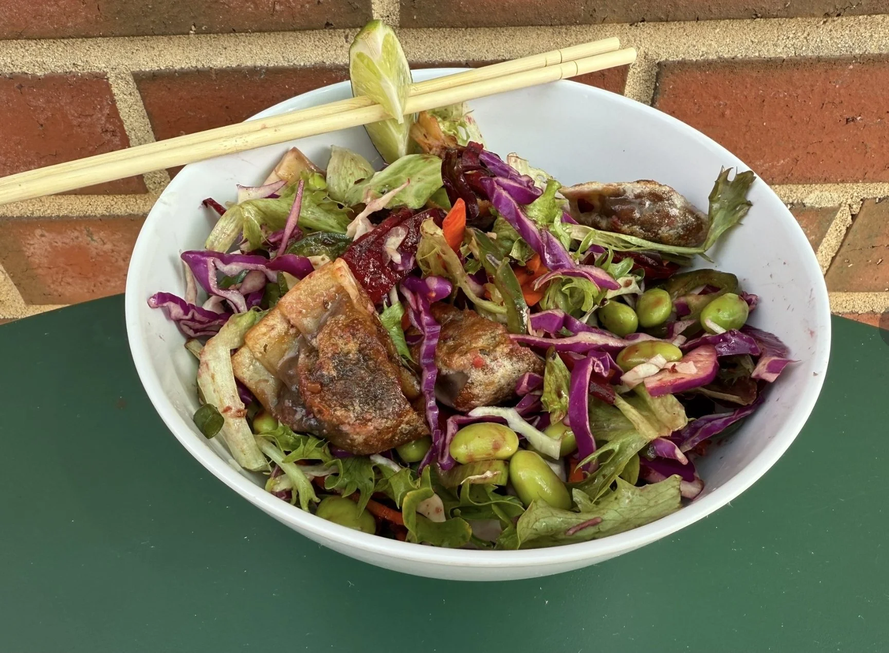 A bowl of mixed salad with purple cabbage, green peas, lettuce, and grilled chicken pieces, placed on a green table with chopsticks on the edge.
