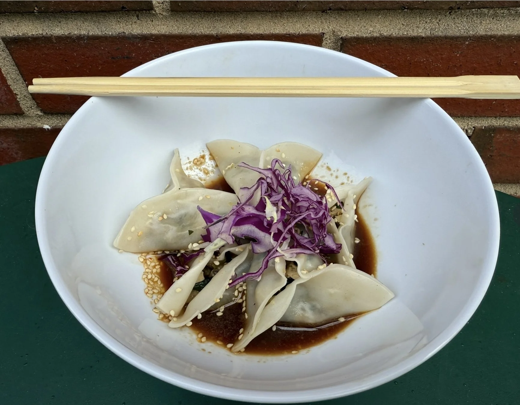 A white bowl containing dumplings topped with shredded purple cabbage, sprinkled with sesame seeds, and drizzled with soy sauce. A pair of wooden chopsticks rests on the bowl's rim, with a brick wall in the background.