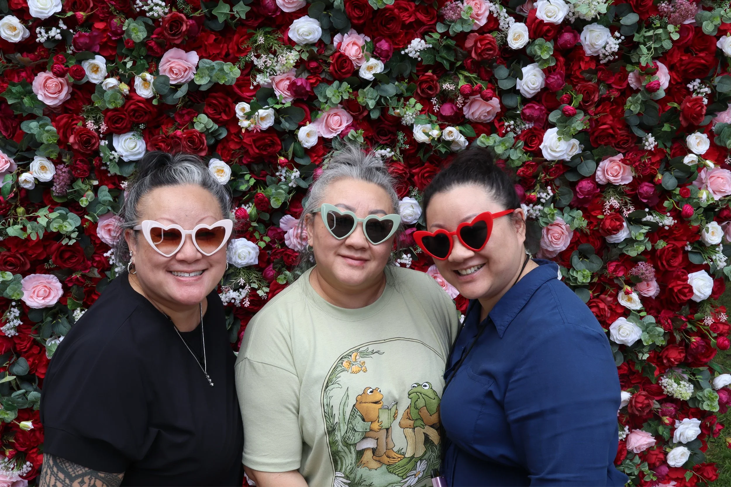 Three women smiling in front of a wall of red, white, and pink flowers, wearing sunglasses with heart-shaped frames.