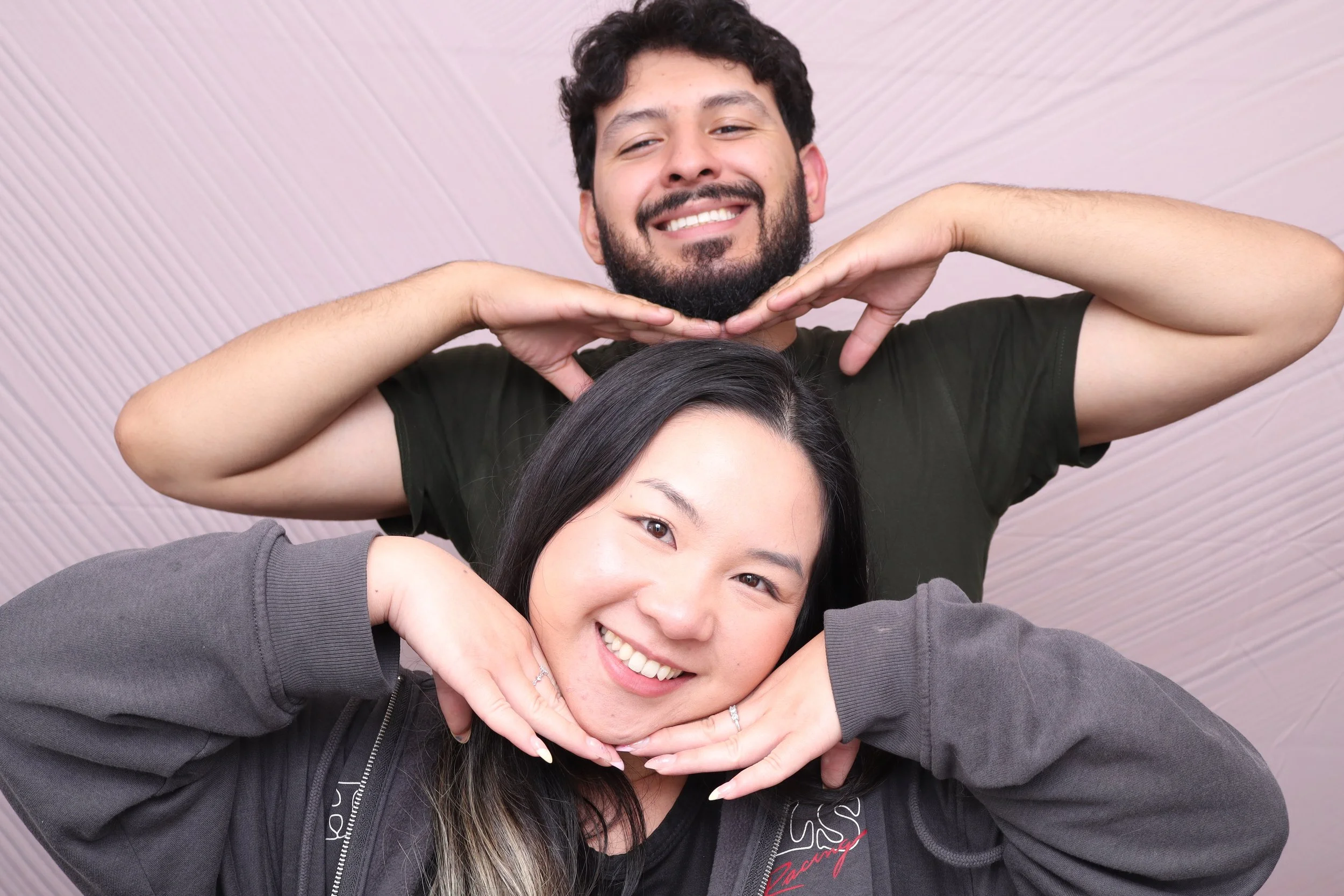 Two smiling people, a woman in front with her hands under her chin, and a man behind with his hands under his chin, making a playful pose.