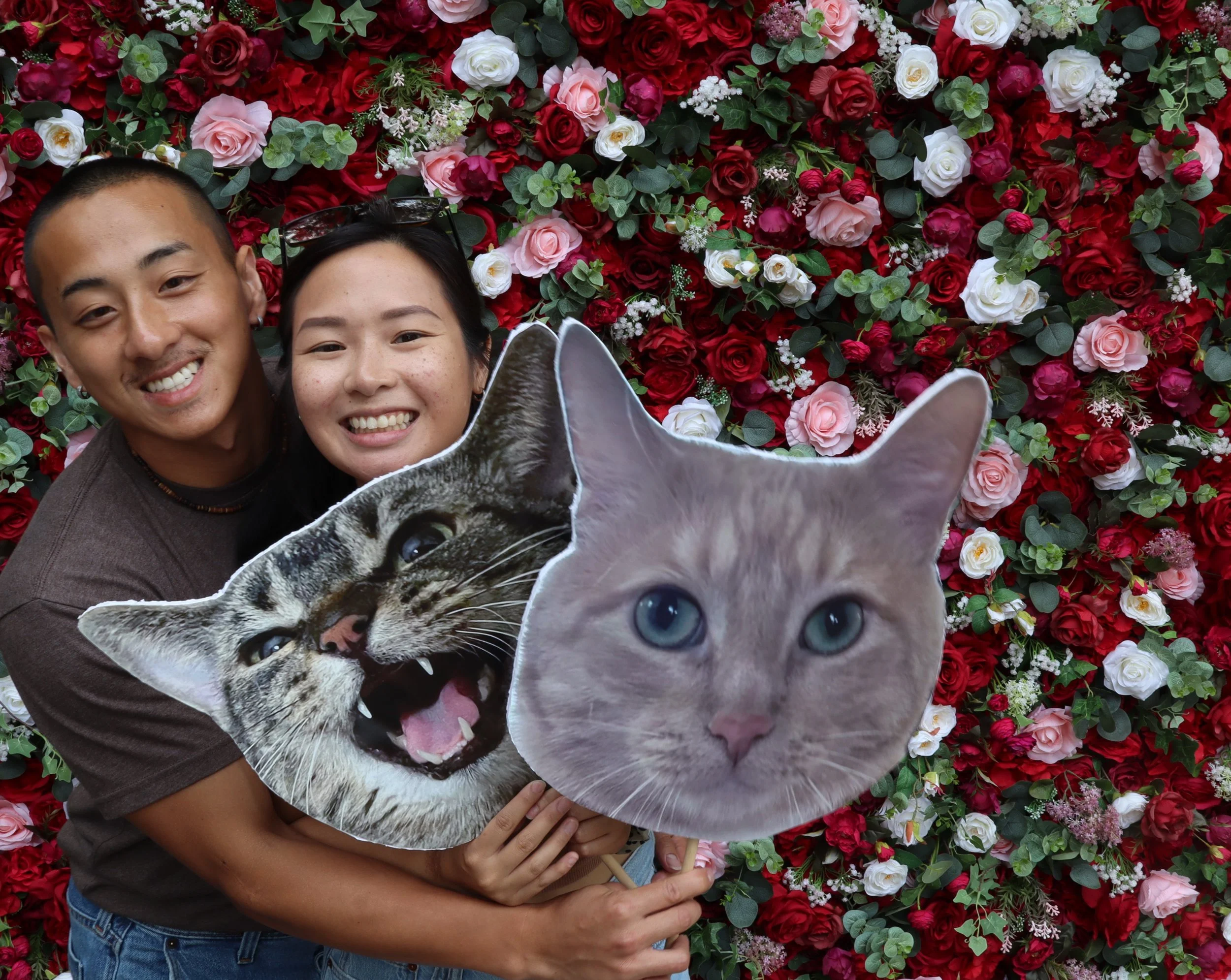 Two smiling people holding cut-out photos of a cat and a tiger in front of a wall of red, pink, and white roses.