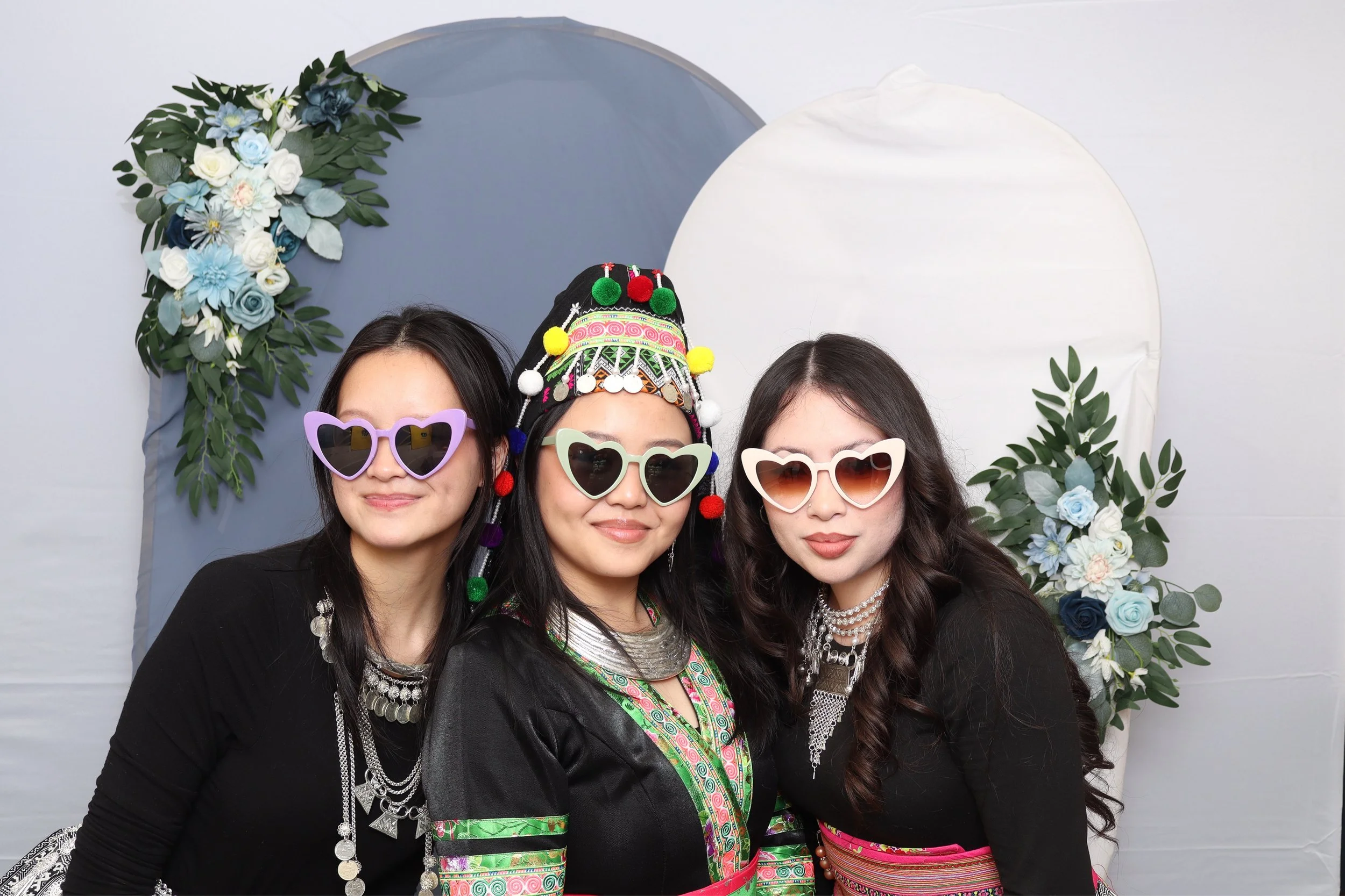 Three women wearing colorful sunglasses and traditional clothing, standing in front of a decorative backdrop with flowers and foliage.