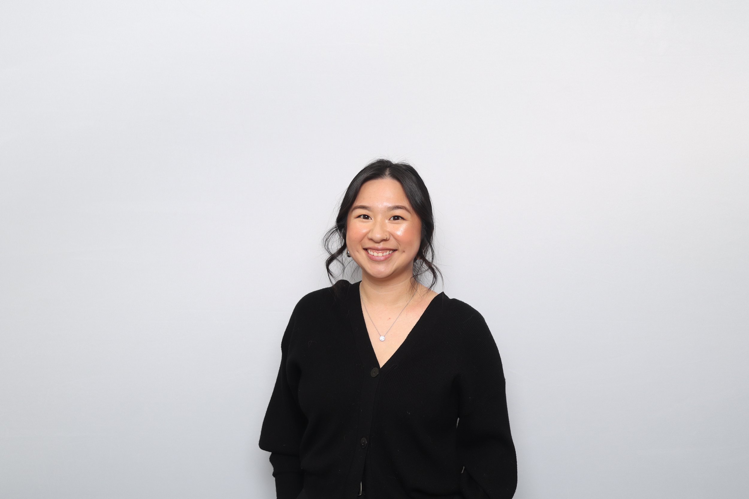 A smiling woman with black hair wearing a black cardigan and necklace, standing against a plain white wall.