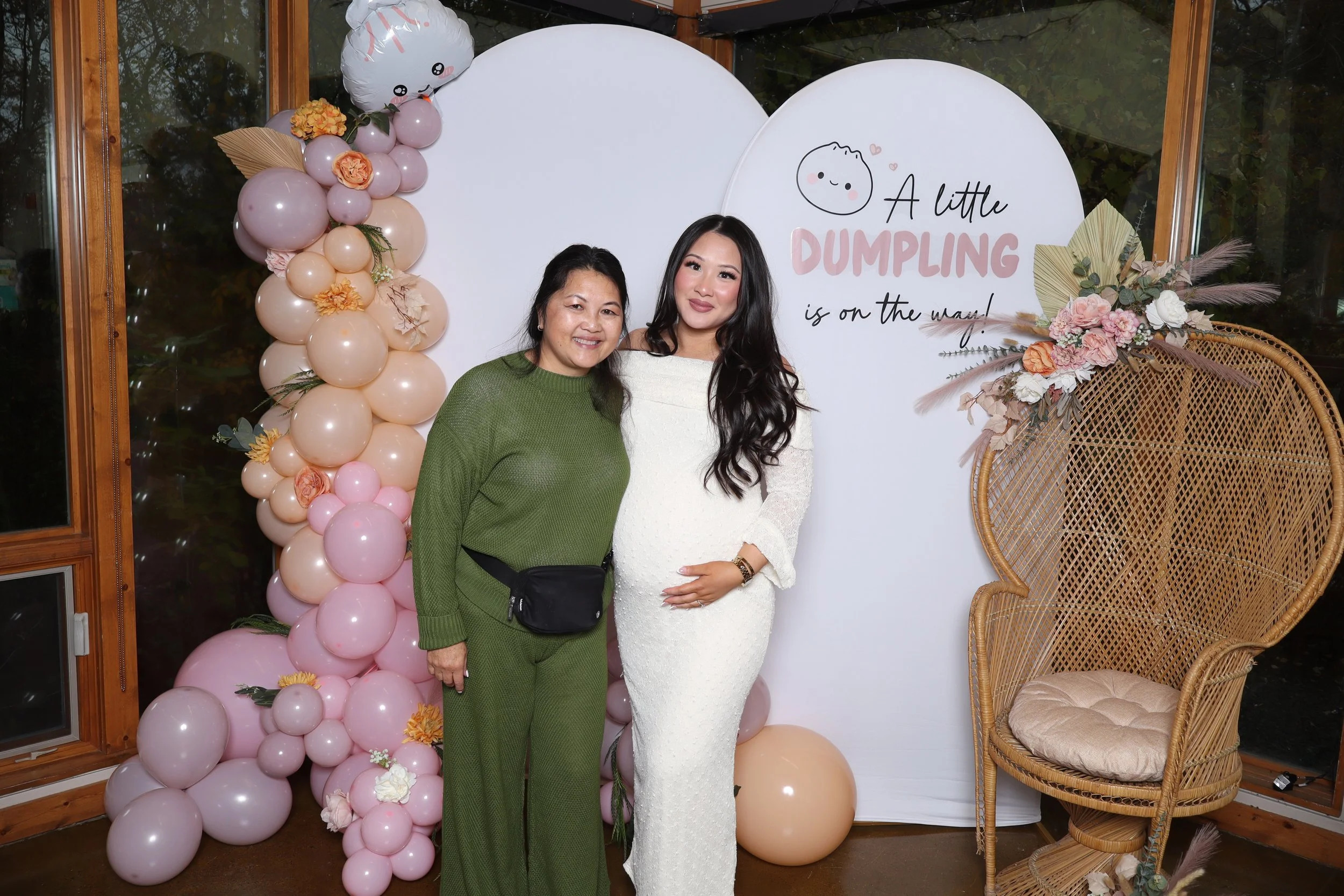 Two women standing in front of a backdrop with balloons and floral decorations. One woman is dressed in a green outfit, and the other in a white dress with a pregnant belly. The backdrop says: "A little DUMPLING is on the way!"
