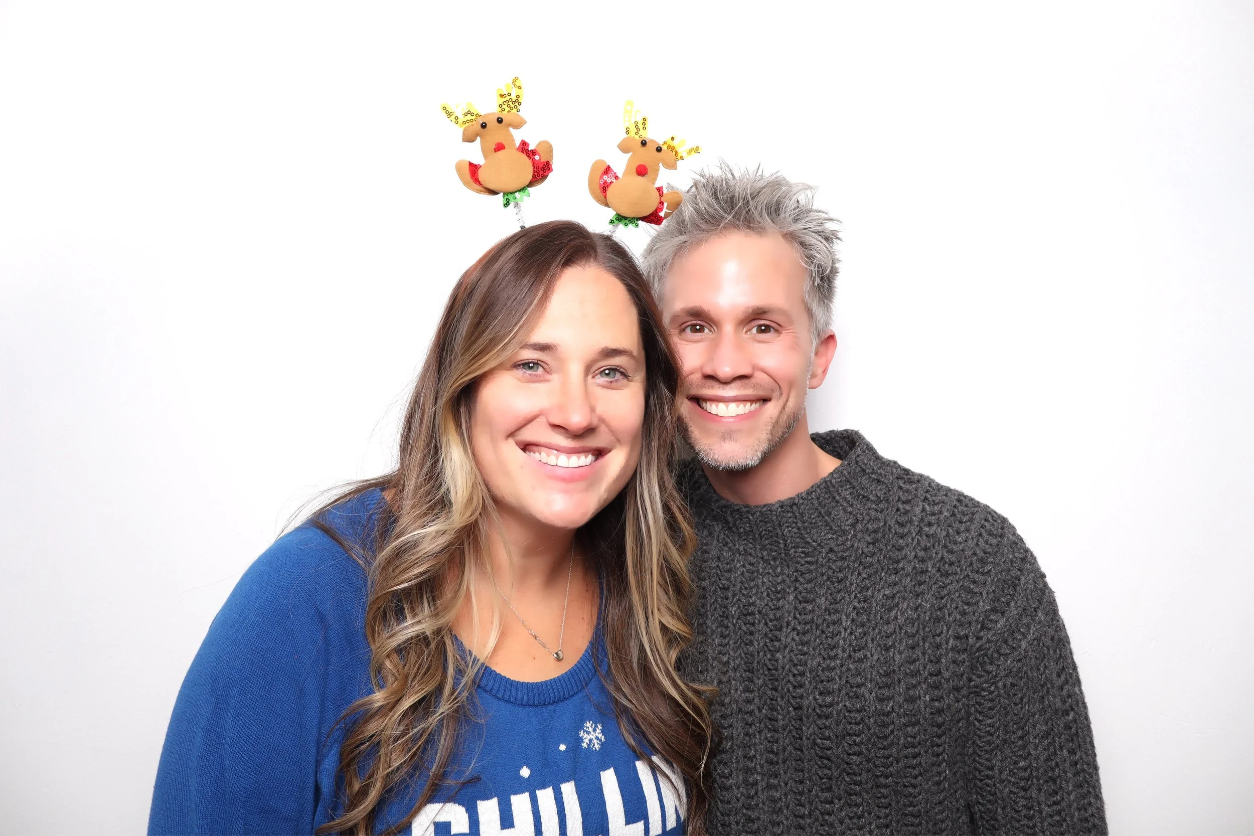 Smiling couple wearing holiday accessories, woman in a blue sweater with snowflakes and the word 'Chill' and man in a gray sweater, standing against a white background.