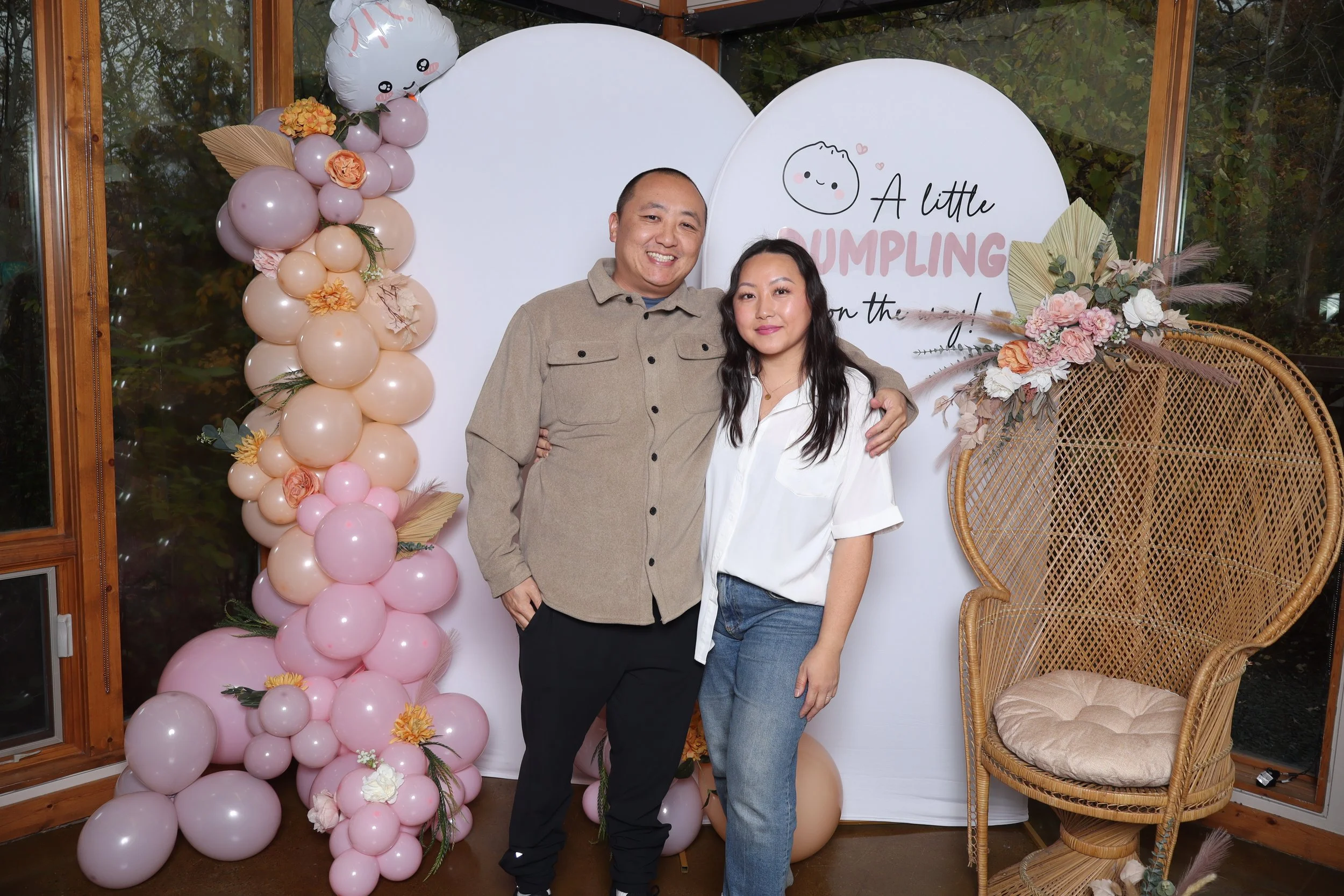 A man and woman standing next to each other, smiling, in front of a decorative background with balloons and floral arrangements, at a celebration event.