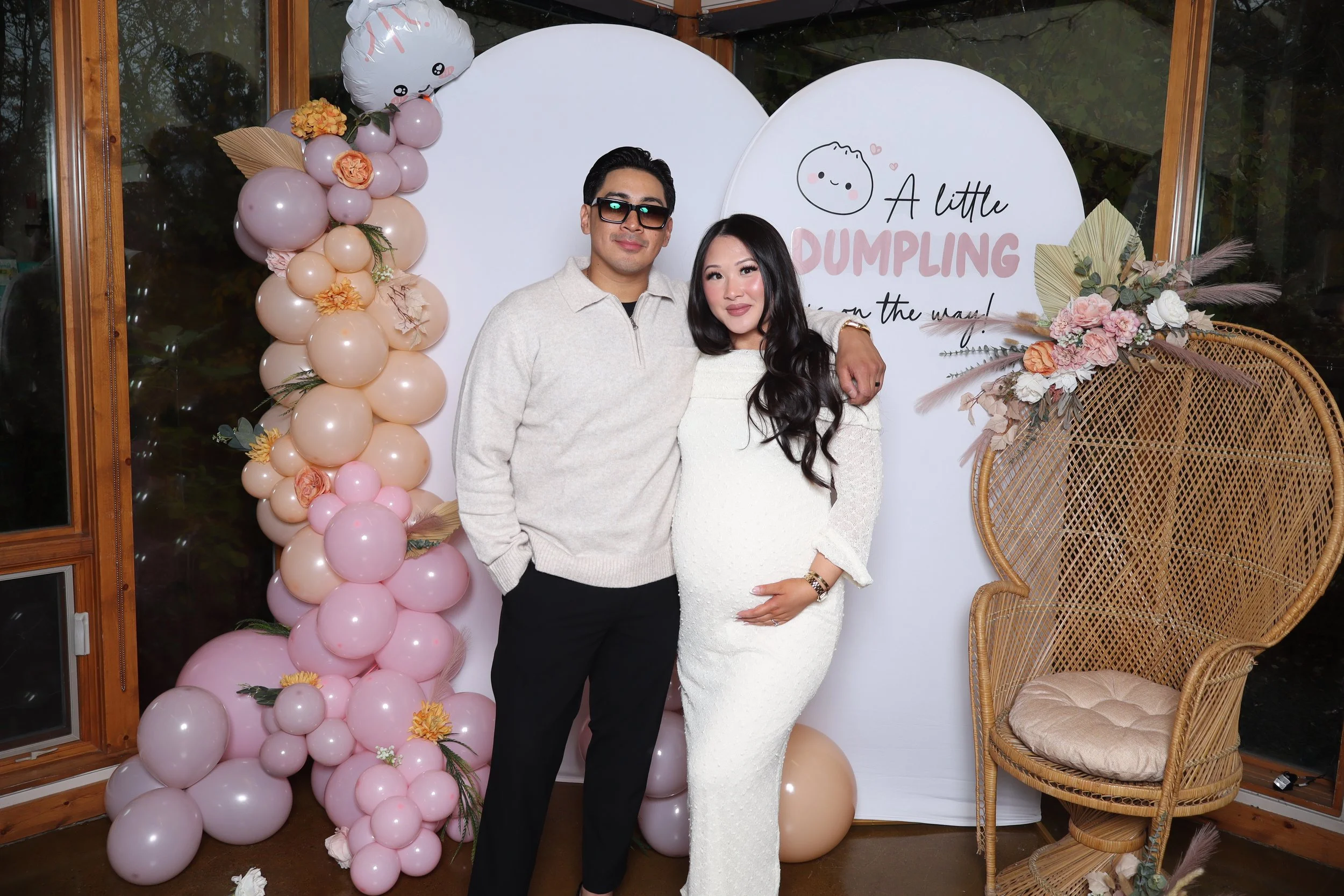 A couple standing in front of a decorated backdrop with balloons and flowers, celebrating a pregnancy or baby shower. The woman is pregnant, wearing a white dress, and the man is wearing a light-colored sweater and sunglasses.