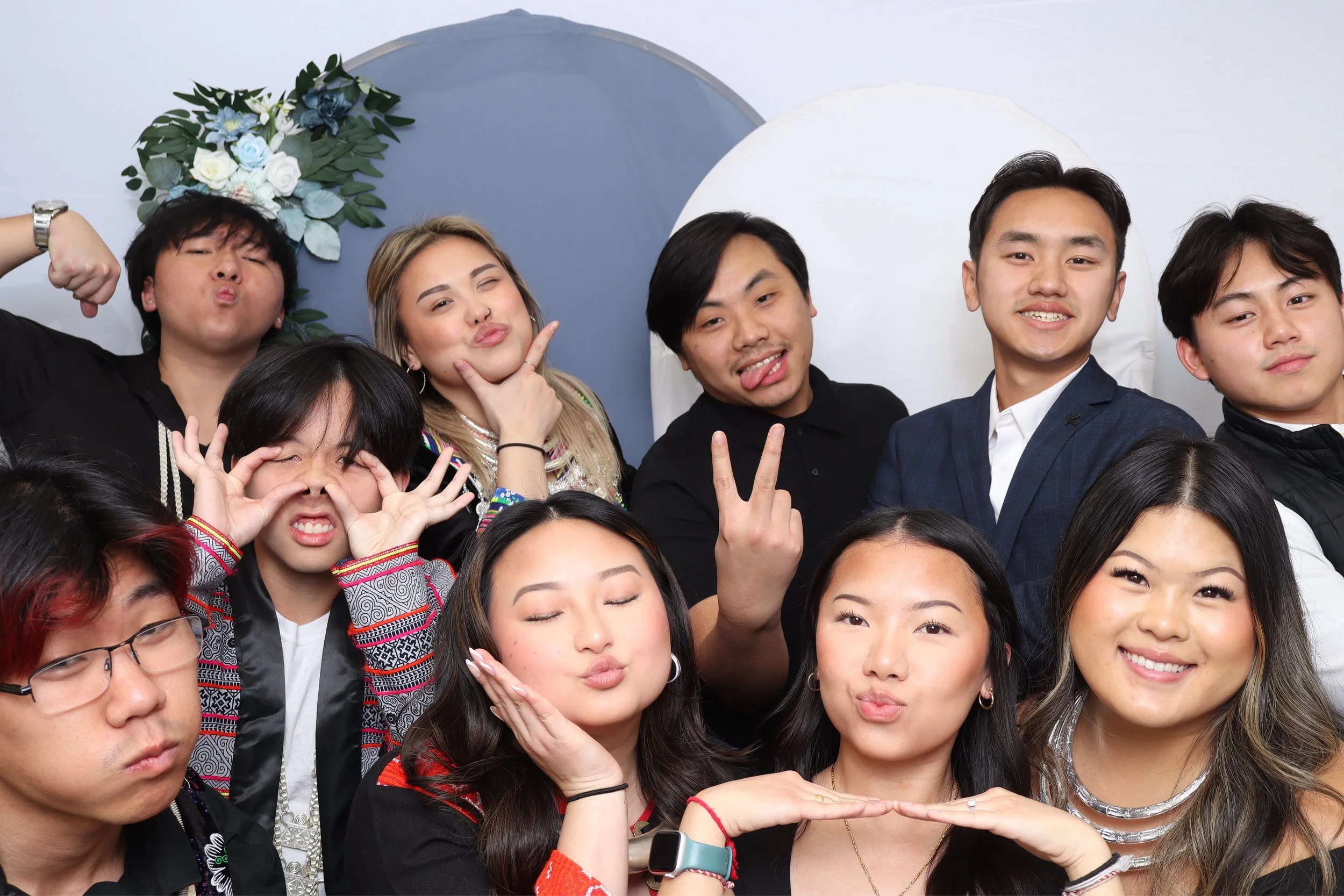 Group of people posing playfully for a photo, some making funny faces and hand gestures, in front of a blue and white backdrop decorated with flowers.
