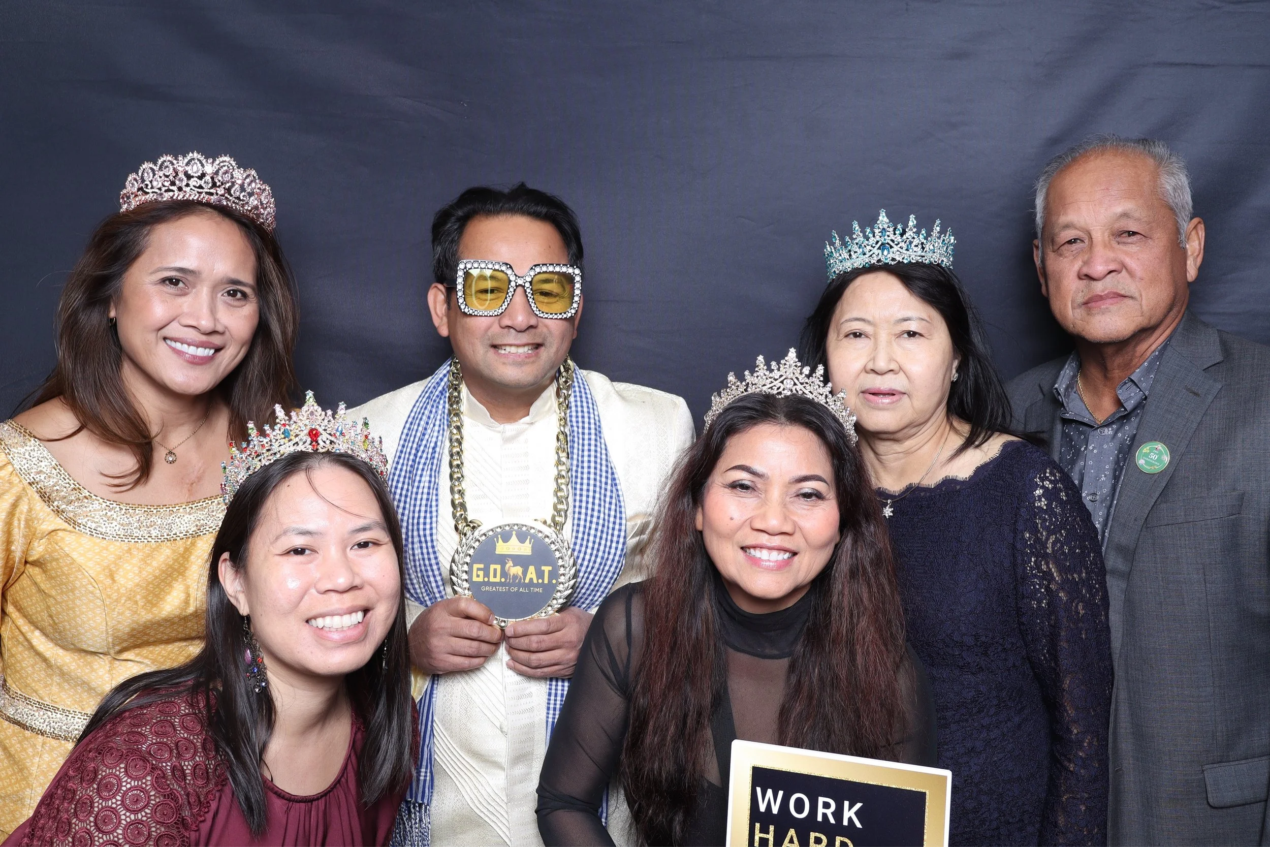 Group of seven people celebrating with crowns and tiaras, dressed in formal and festive attire, holding a sign that reads 'WORK HARD, BE NICE' in a photo booth with a dark background.