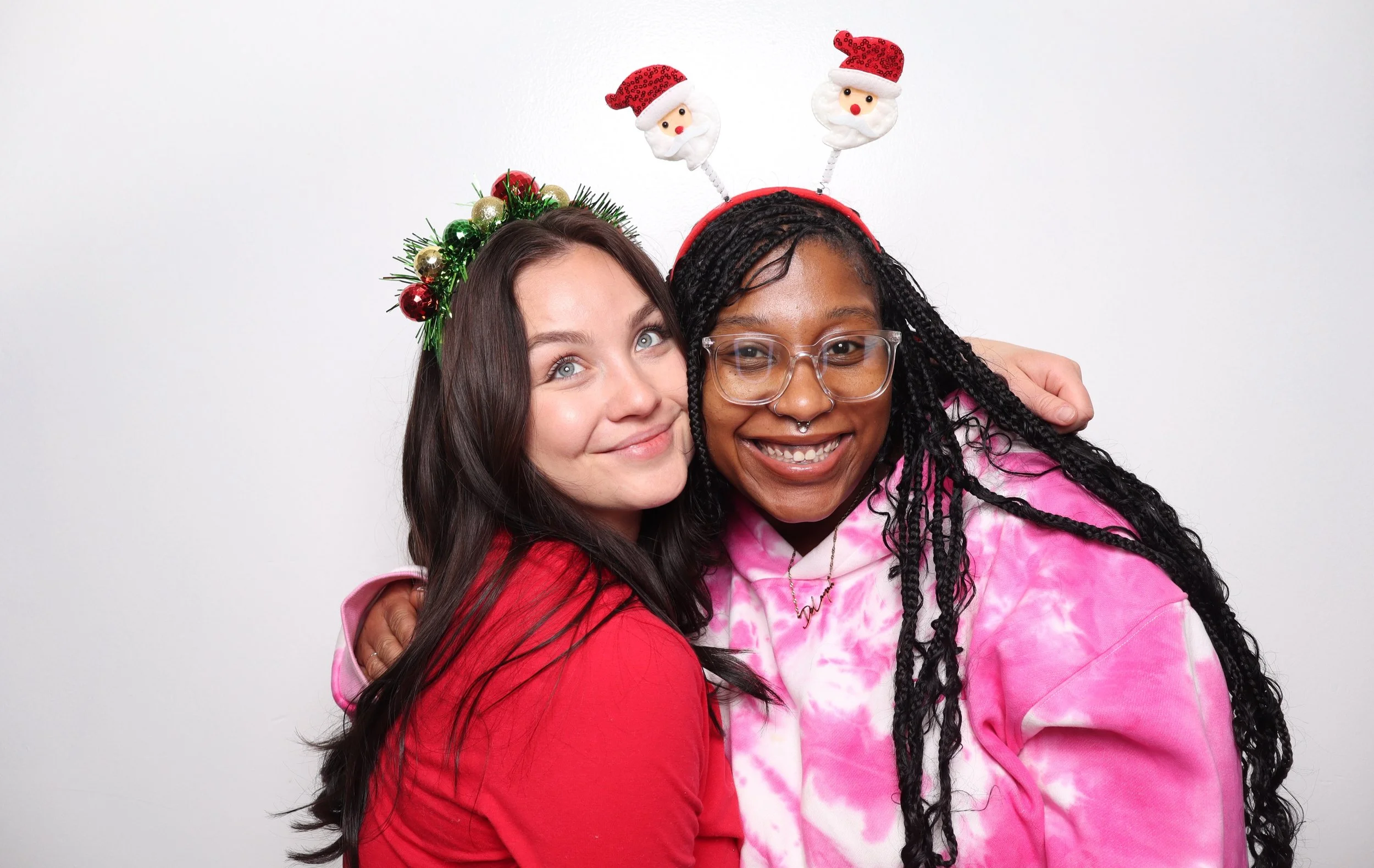 Two women wearing Christmas-themed accessories smiling and hugging against a plain white background. One has a headband with Christmas ornaments, and the other has a headband with Santa face and reindeer antlers.