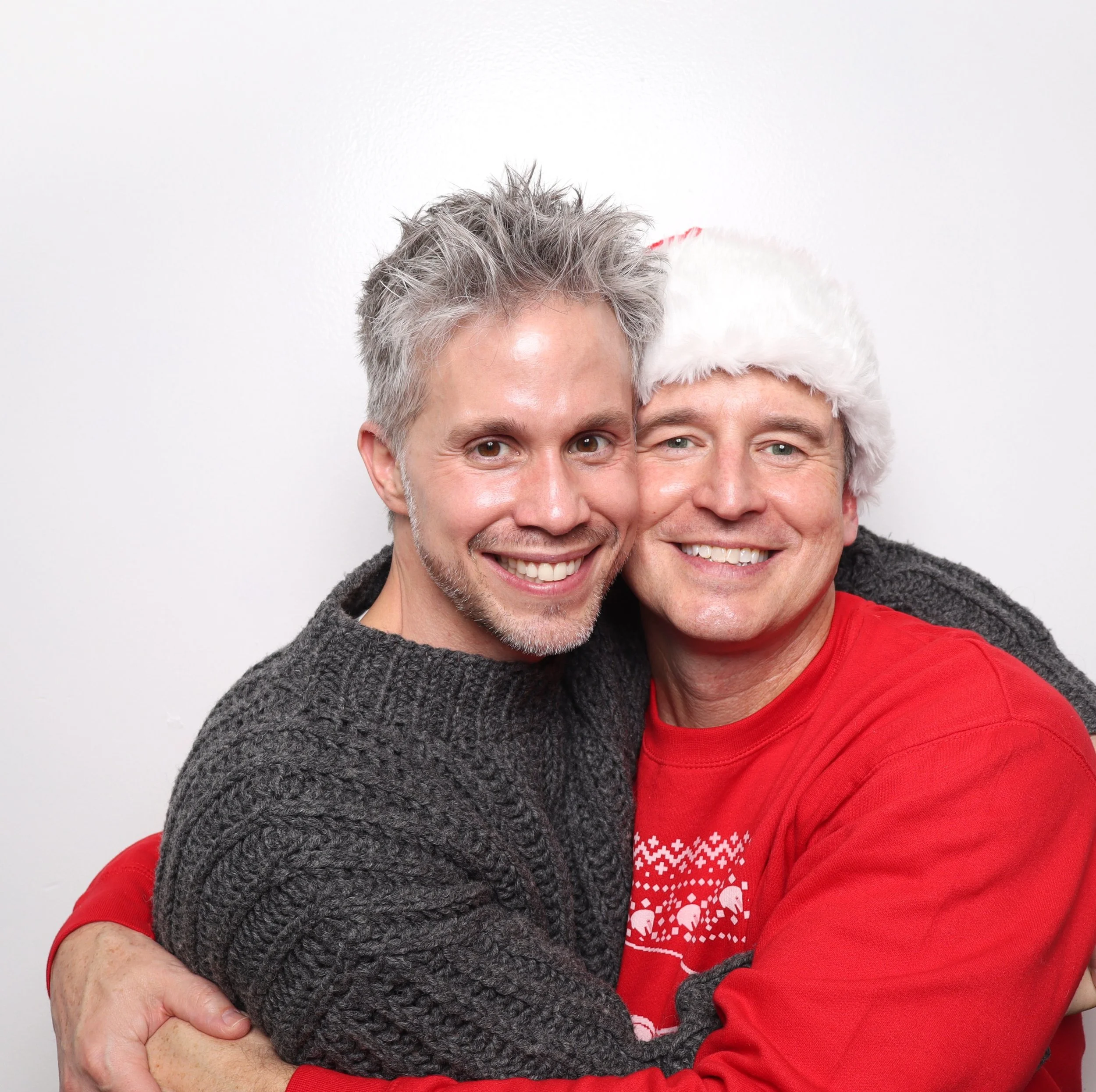 Two smiling men hugging, one wearing a Santa hat and a red Christmas sweater, the other in a gray knitted sweater, against a plain white background.