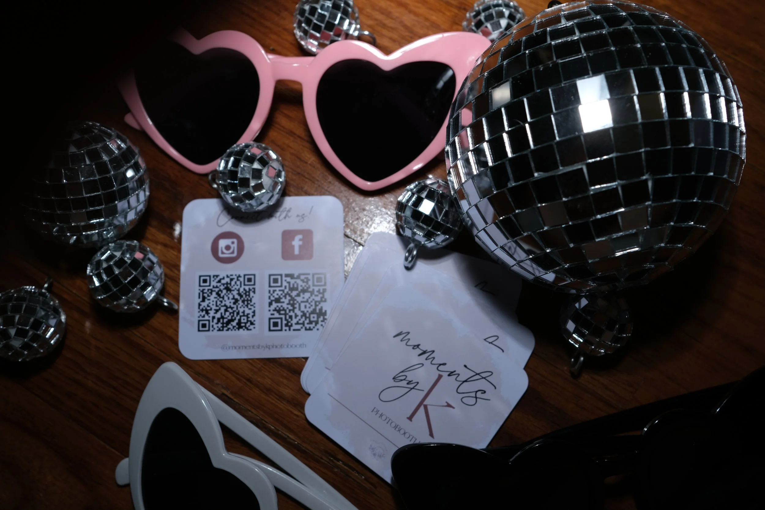 Pink heart-shaped sunglasses, multiple small and large disco balls, business cards, and social media QR codes arranged on a wooden surface.