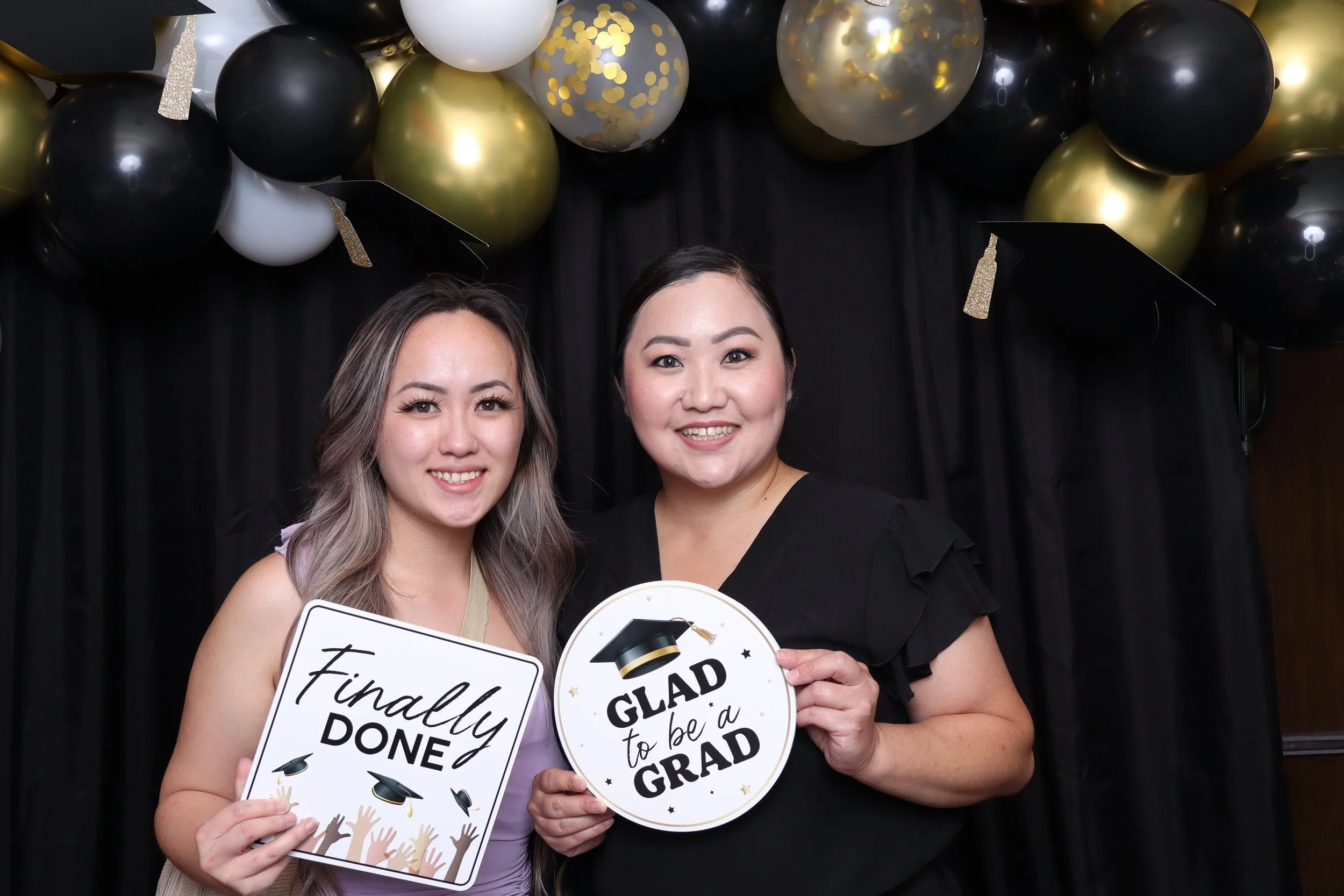 Two women celebrating graduation, one holding a sign that says "Finally Done" and the other holding a sign that says "Glad to be a Grad," with black, white, and gold balloon decorations in the background.