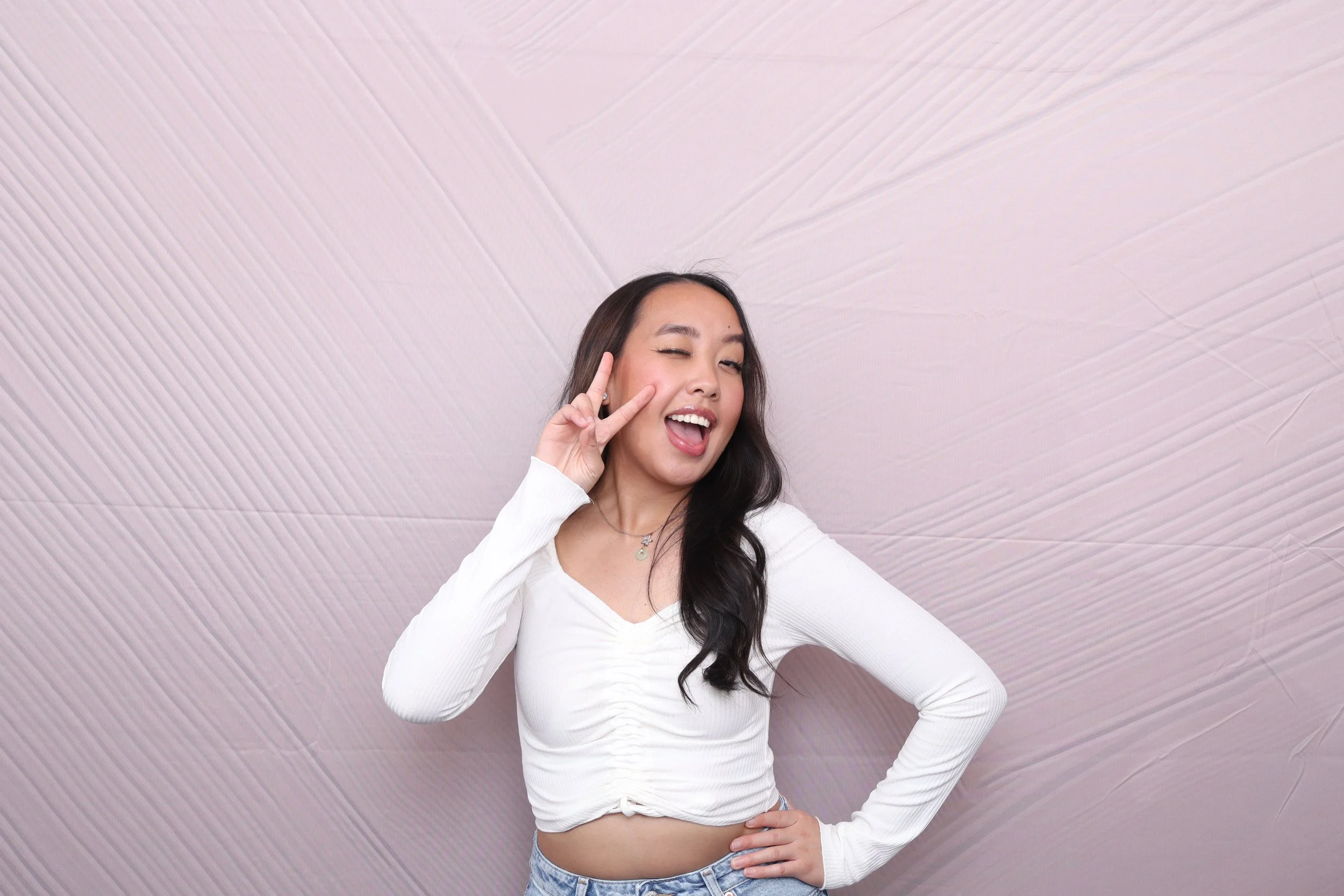 A young woman with long dark hair making a peace sign near her eye, winking and smiling, with a playful expression, standing against a textured pink wall. She is wearing a white long-sleeve top and light blue jeans.