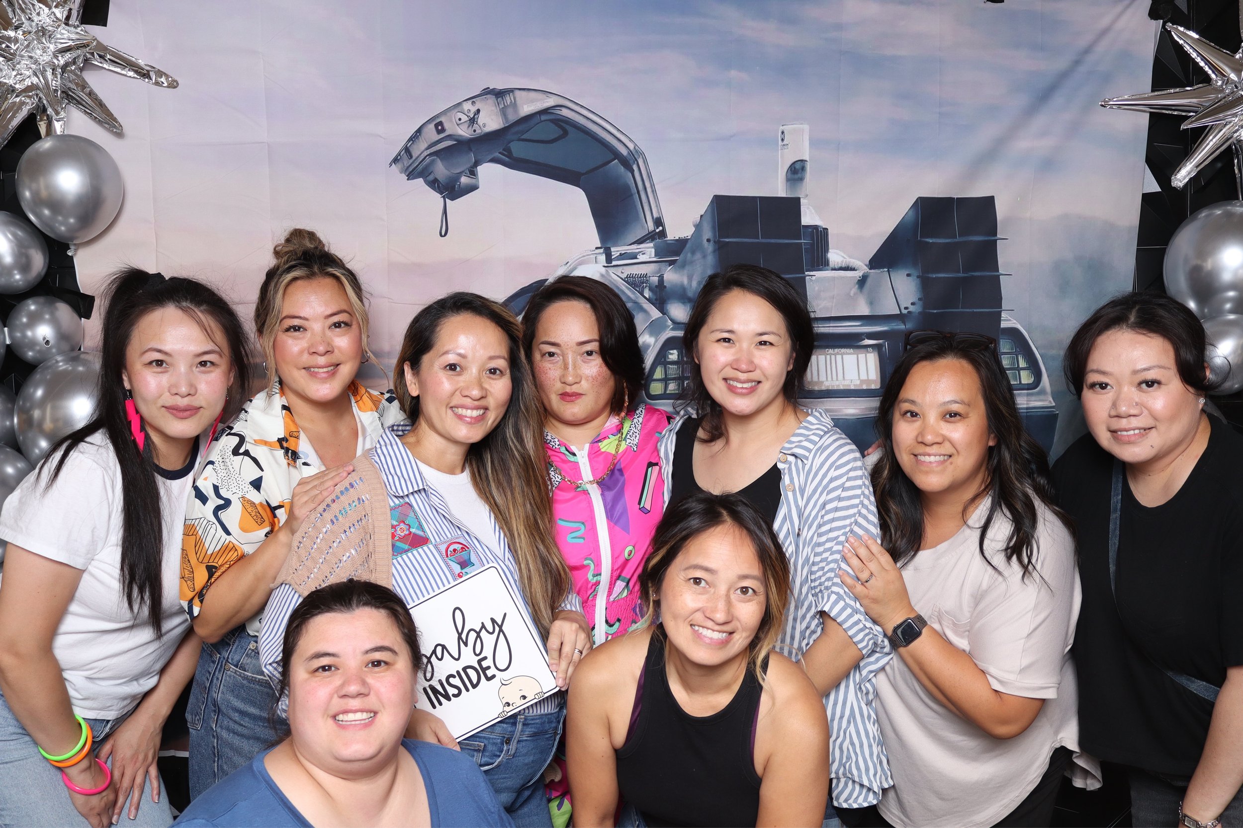 Group of nine women posing together indoors with a space-themed backdrop and silver balloons.