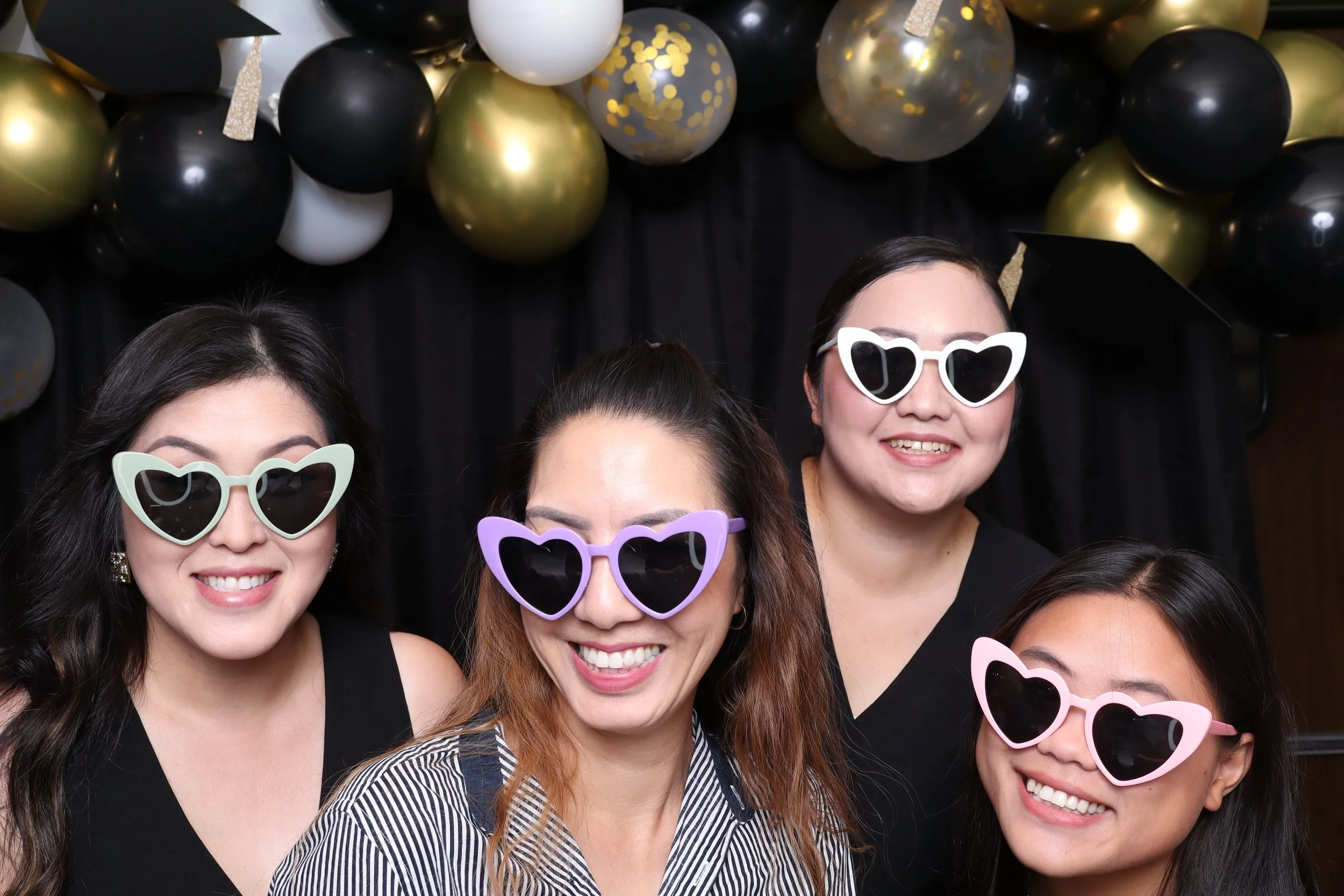 Four women at a celebration wearing colorful heart-shaped sunglasses, smiling, with black, gold, and gray balloons decorating the background.