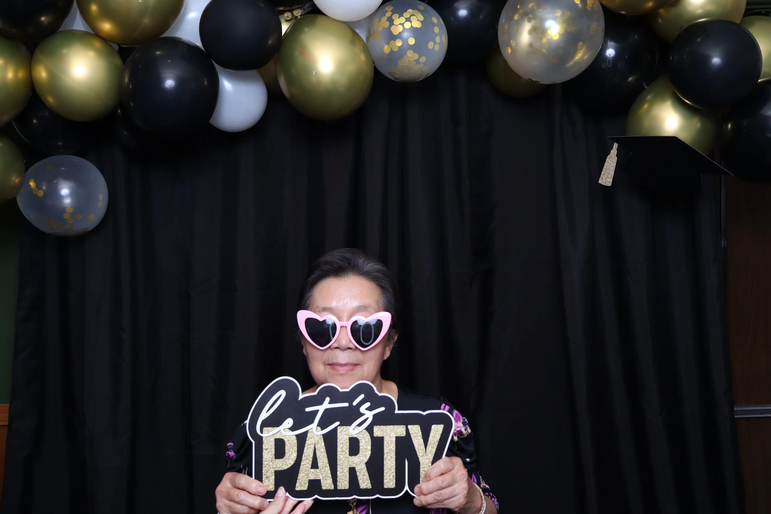 An older woman wearing heart-shaped sunglasses holding a sign that says "let's PARTY" with a black curtain backdrop and balloon decorations in gold, black, white, and clear with gold confetti.