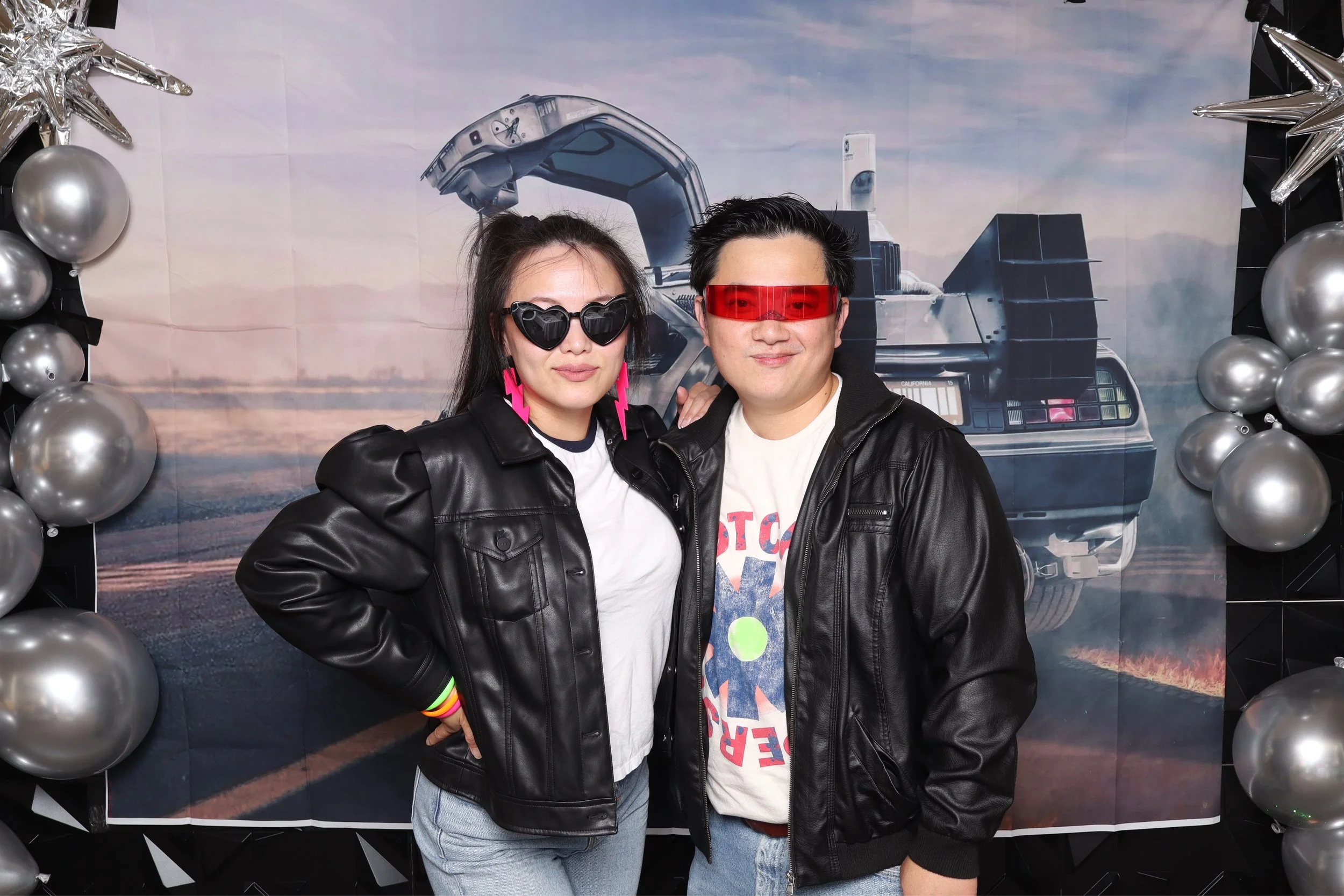 Two people dressed in retro-futuristic costumes posing in front of a backdrop featuring the DeLorean time machine from the movie Back to the Future, surrounded by silver balloons and star-shaped decorations.