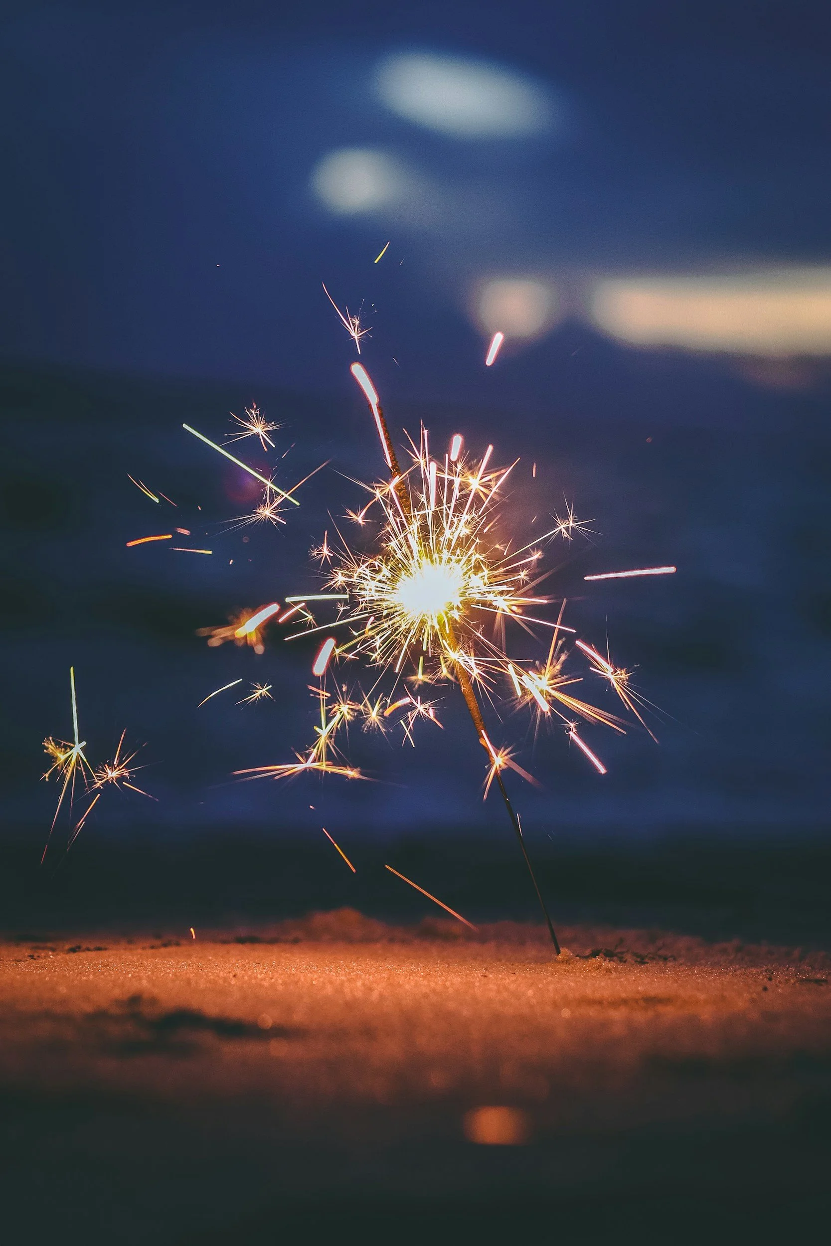 A lit sparkler burning on a dark surface with sparks flying outward against a dark blue sky with clouds.