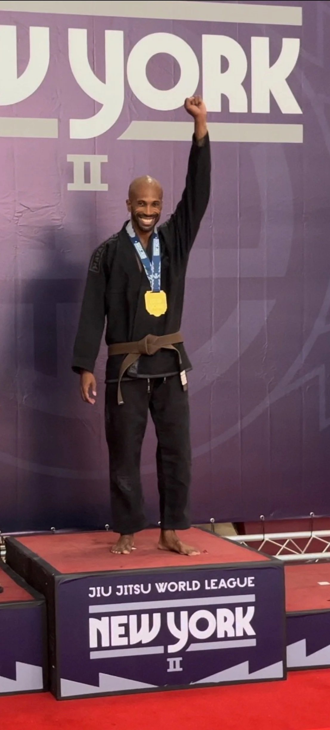 A man in a black martial arts gi with a brown belt, barefoot, standing on a winner's podium, raising his right hand in victory, wearing a medal around his neck, at a Brazilian Jiu Jitsu event in New York.