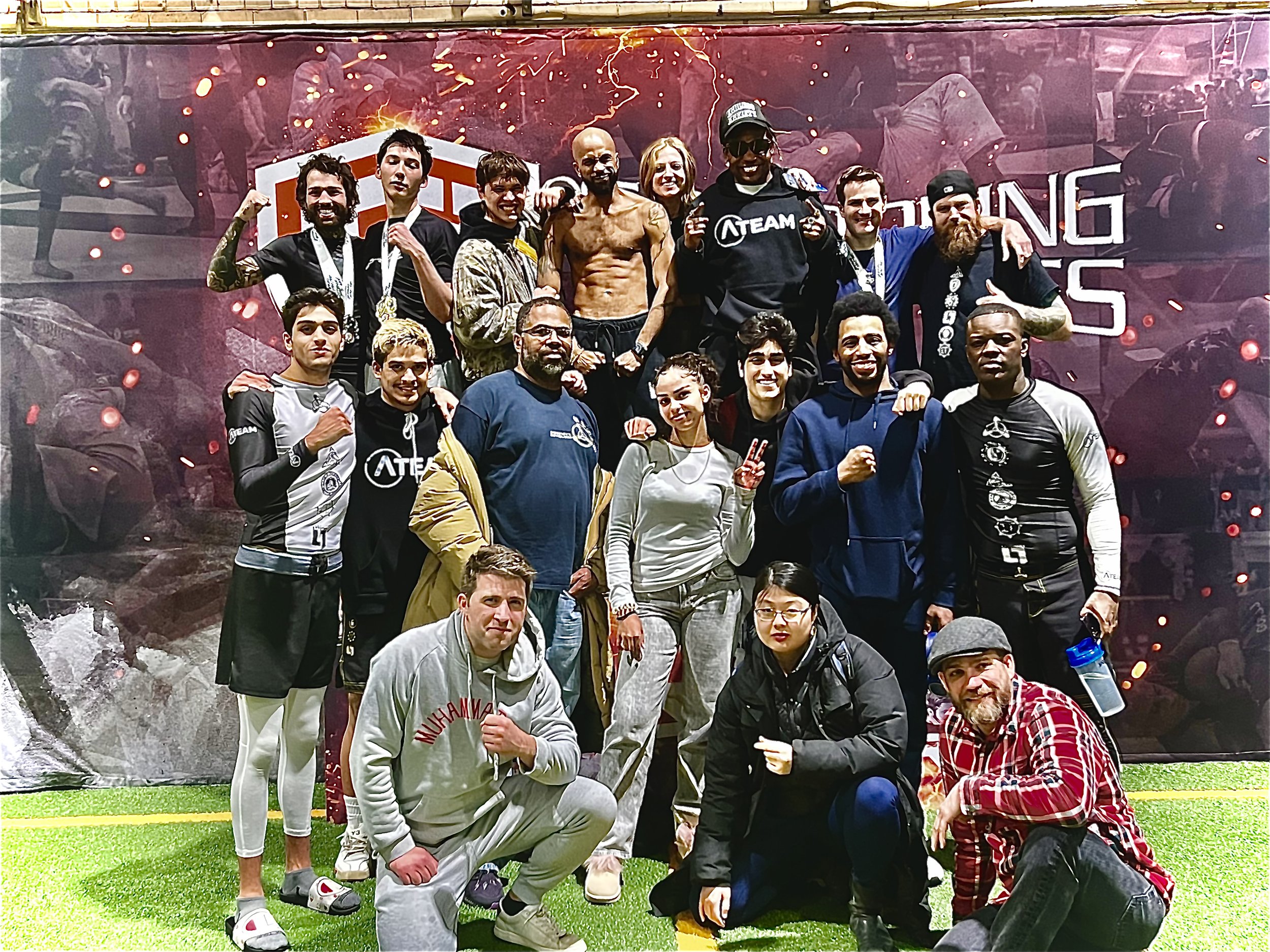 A group of diverse people, some in athletic gear, posing together on a grassy indoor field after participating in a grappling tournament, with a large backdrop displaying graphics and text.
