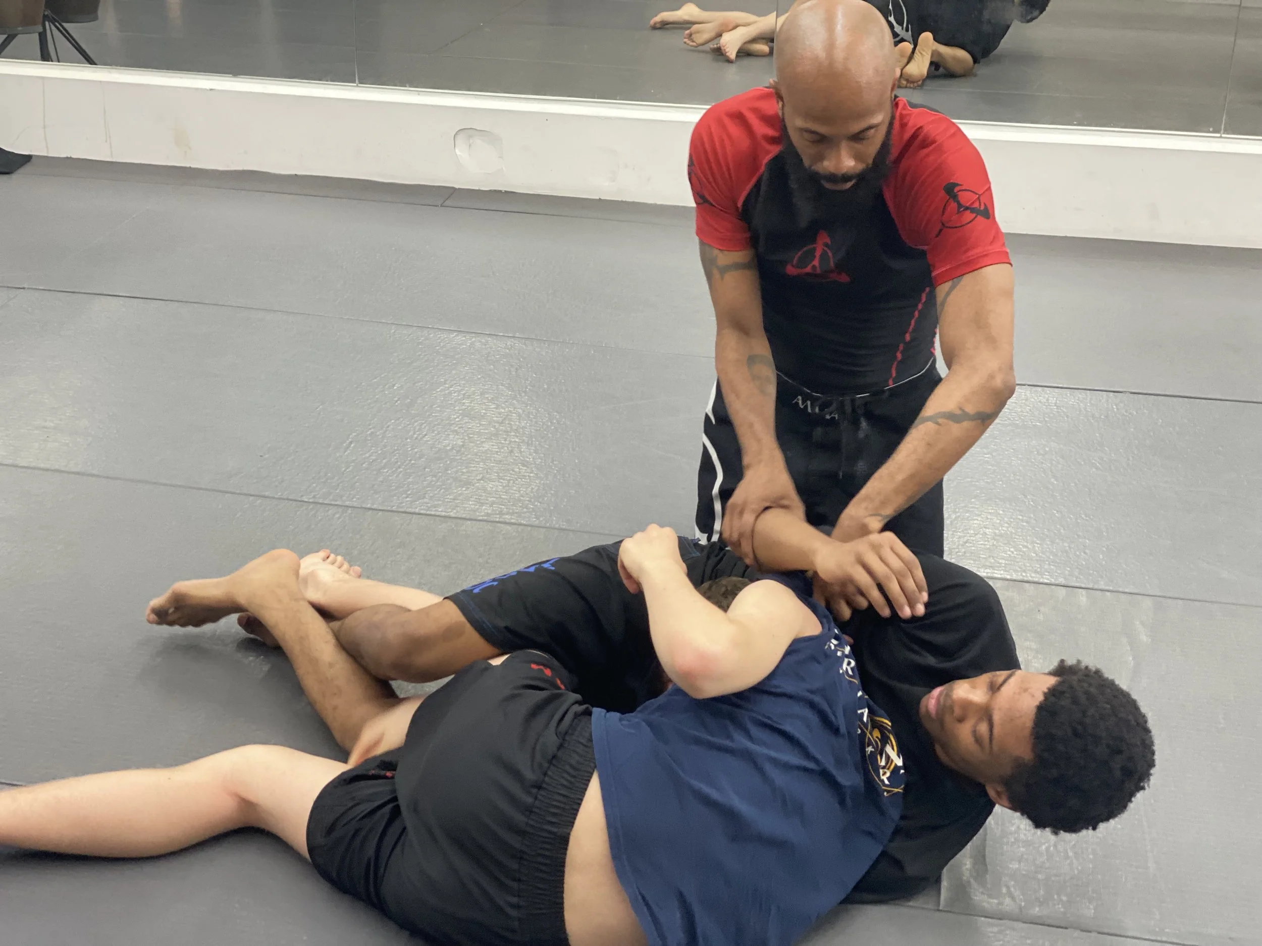 Two men practicing Brazilian Jiu-Jitsu on a gray mat; one in black shorts and blue shirt, the other in black shorts and black shirt, with an instructor in a red and black rash guard demonstrating a chokehold.