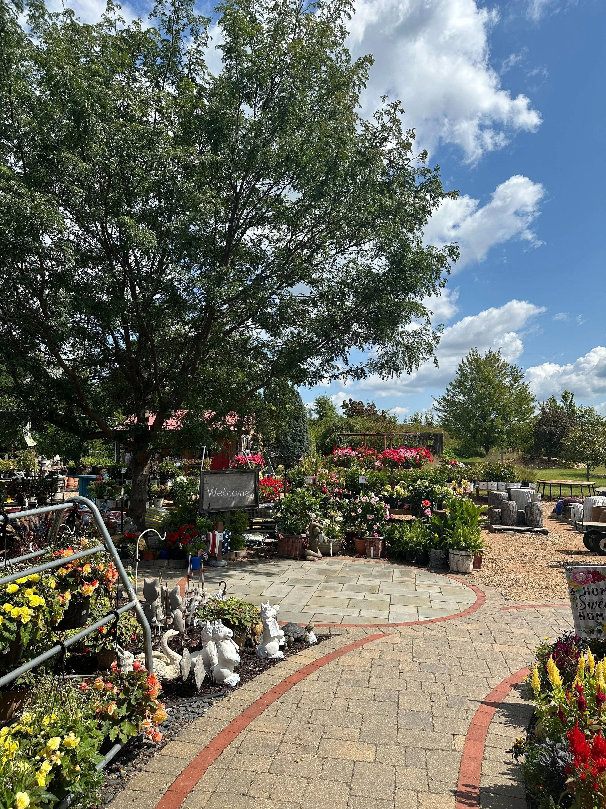 A garden center with colorful flowers, trees, gardening accessories, and a welcoming sign under a partly cloudy sky.