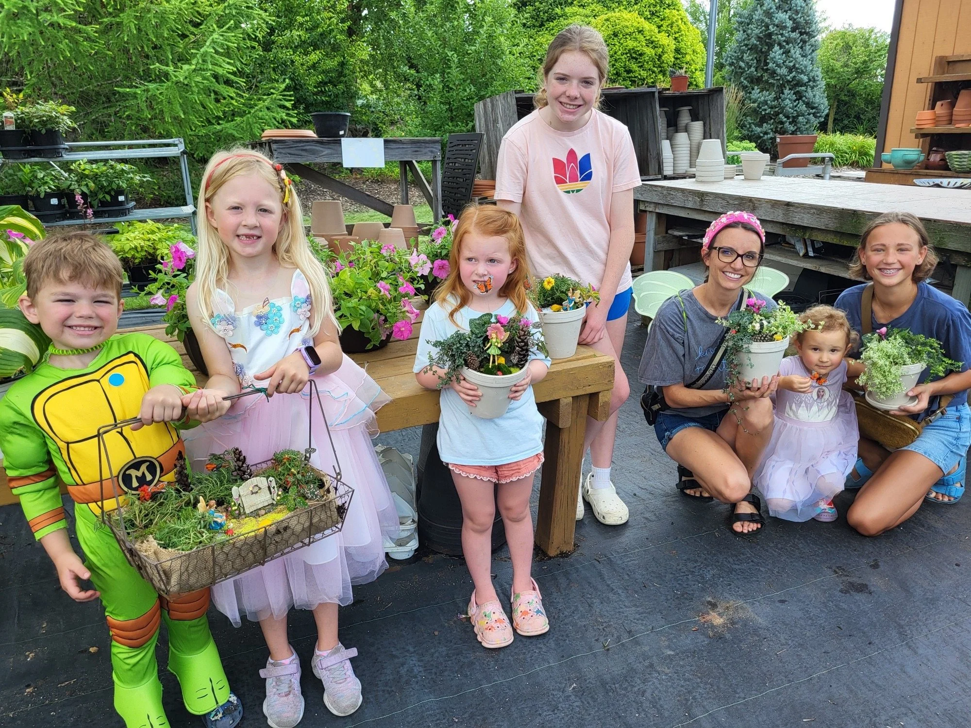 A group of children and a woman with glasses holding various potted plants and flowers, posing outdoors at a plant nursery or garden center. The children are dressed in costumes and casual clothes, smiling.