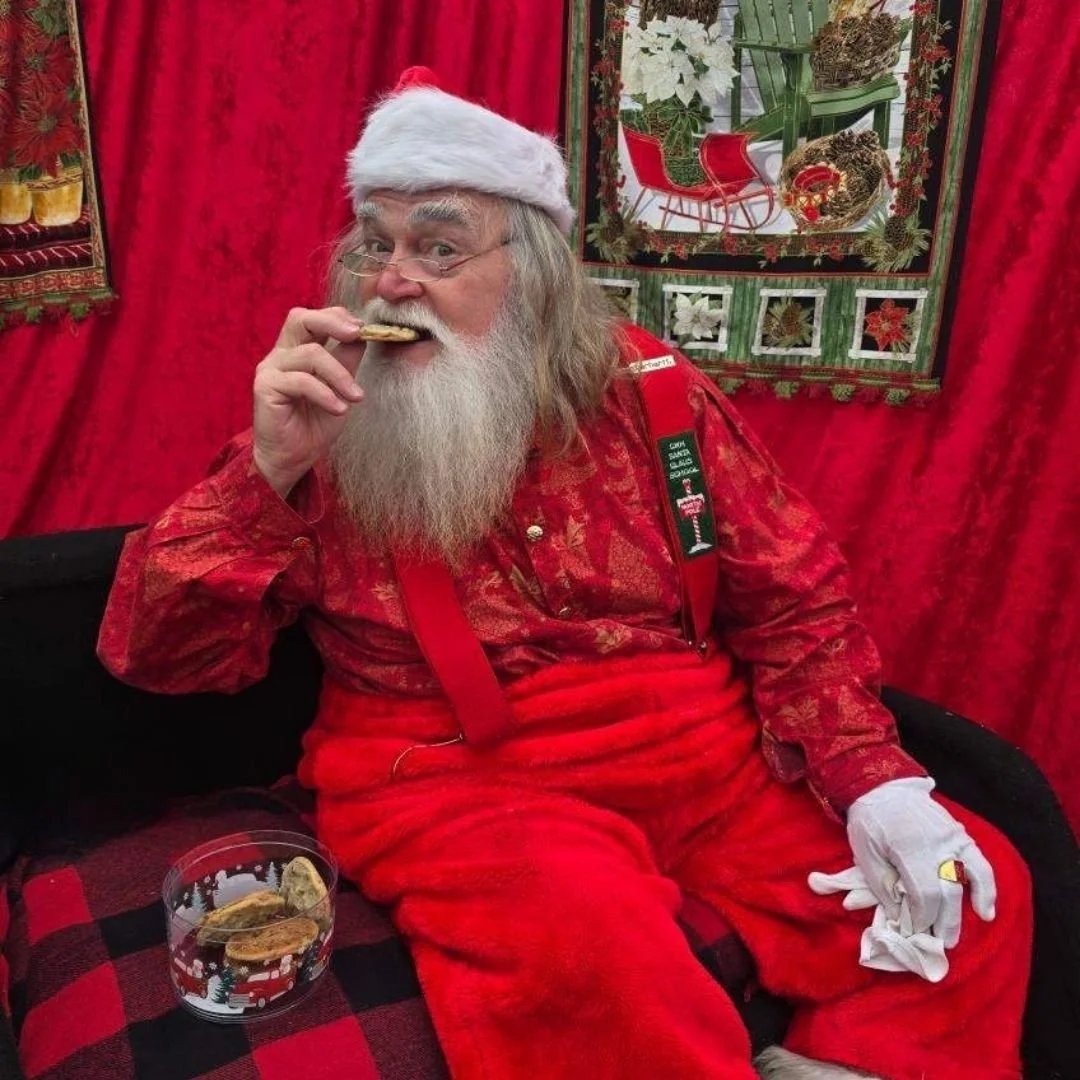 A man dressed as Santa Claus, with a long grey beard and wearing a red outfit and Santa hat, is sitting on a black and red plaid couch. He is holding a cookie near his mouth, and there is a glass bowl of cookies on his lap. The background features red curtains and Christmas-themed decorations.