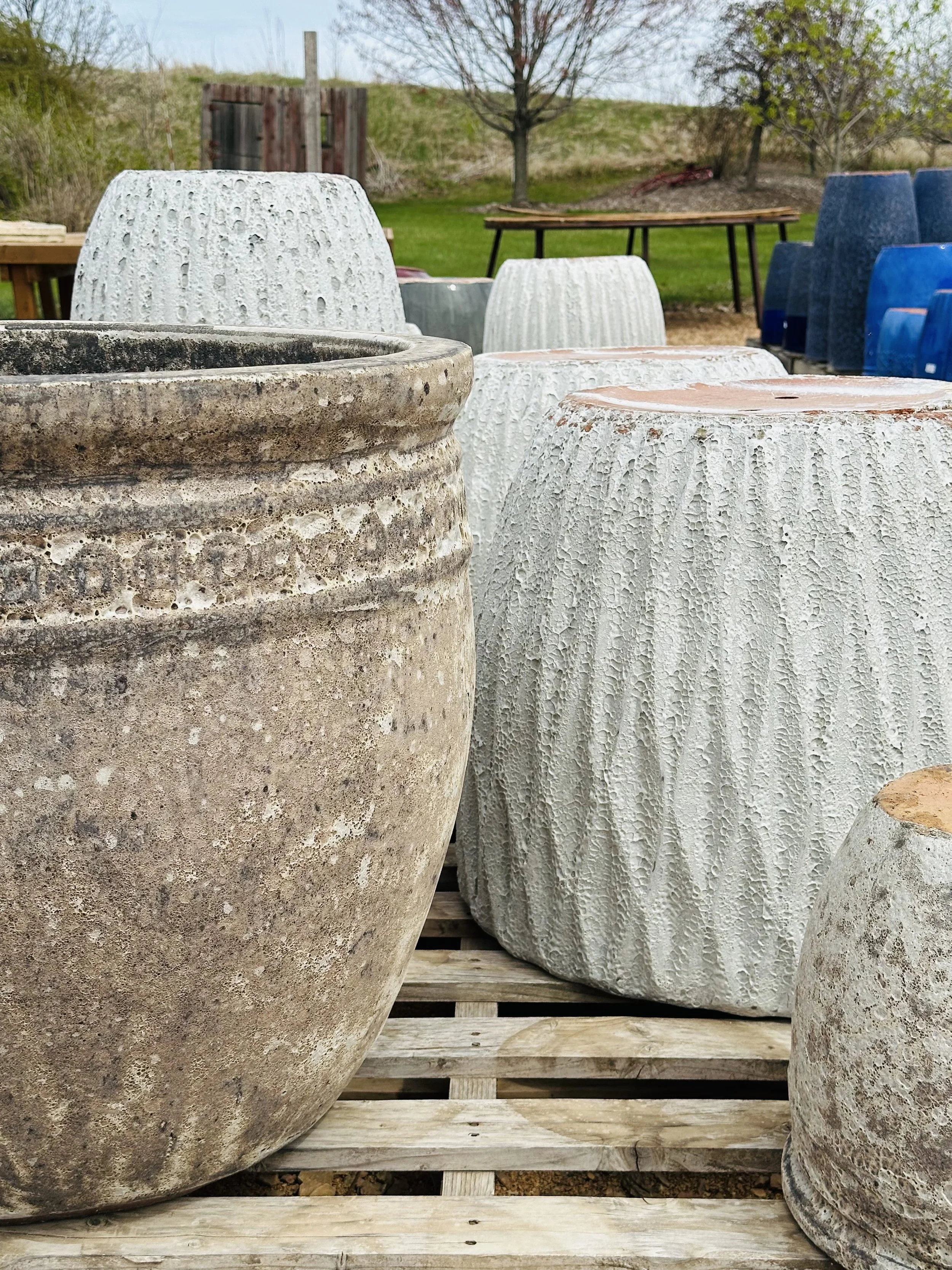 Various textured and colored outdoor planters arranged on a wooden pallet with a lawn, trees, and a fence in the background.