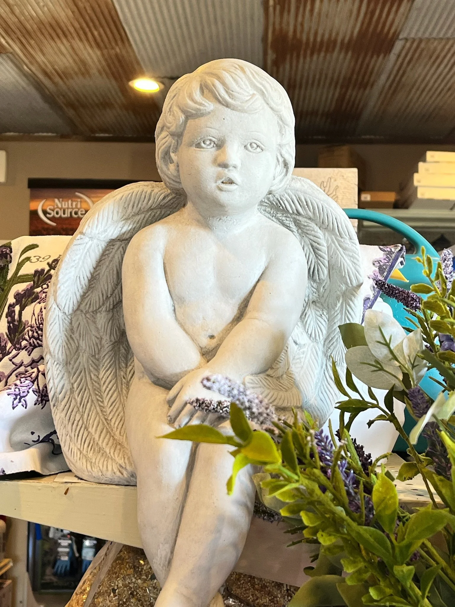 White angel statue with childlike face, seated on a surface with arms crossed and resting on one knee, surrounded by artificial flowers.