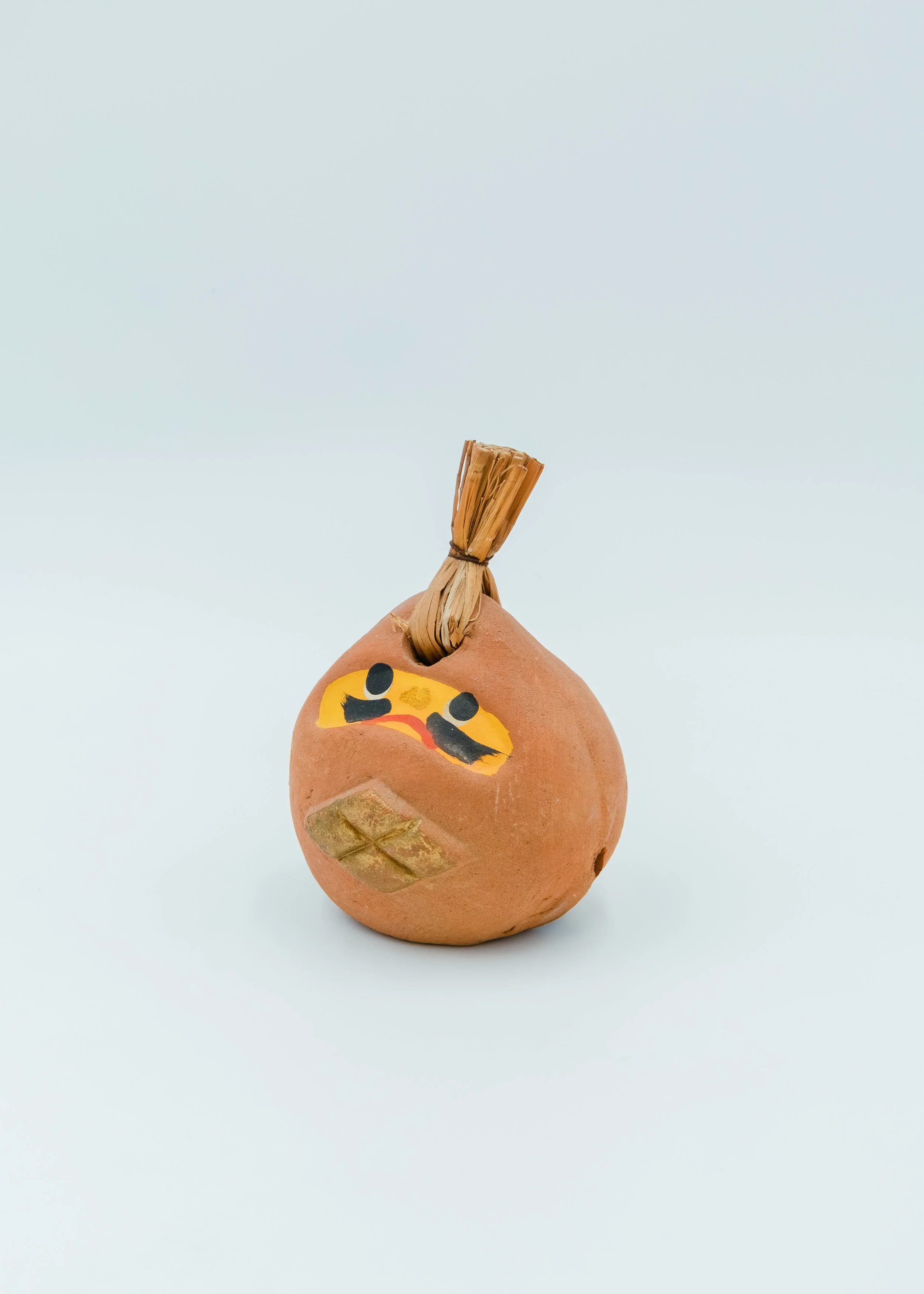 Three-quarter view of UNGLAZED TALISMAN 1970, a rare vintage Japanese earthen bell (Dorei) with hand-painted Daruma face and a traditional straw bundle top.
