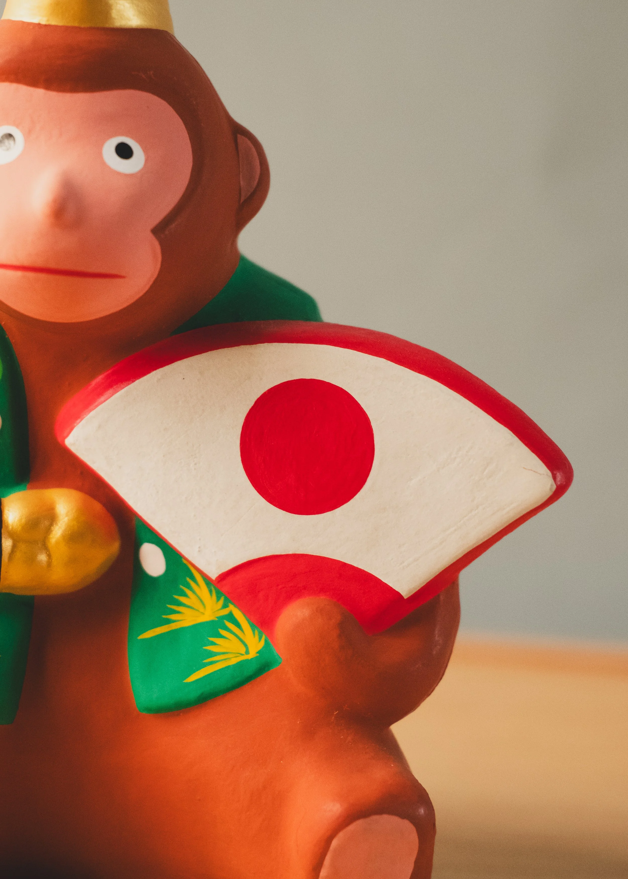 Detailed shot of the Hinomaru rising sun fan held by the Hanamaki monkey. A spiritual utility symbolizing forward momentum and rising fortune.