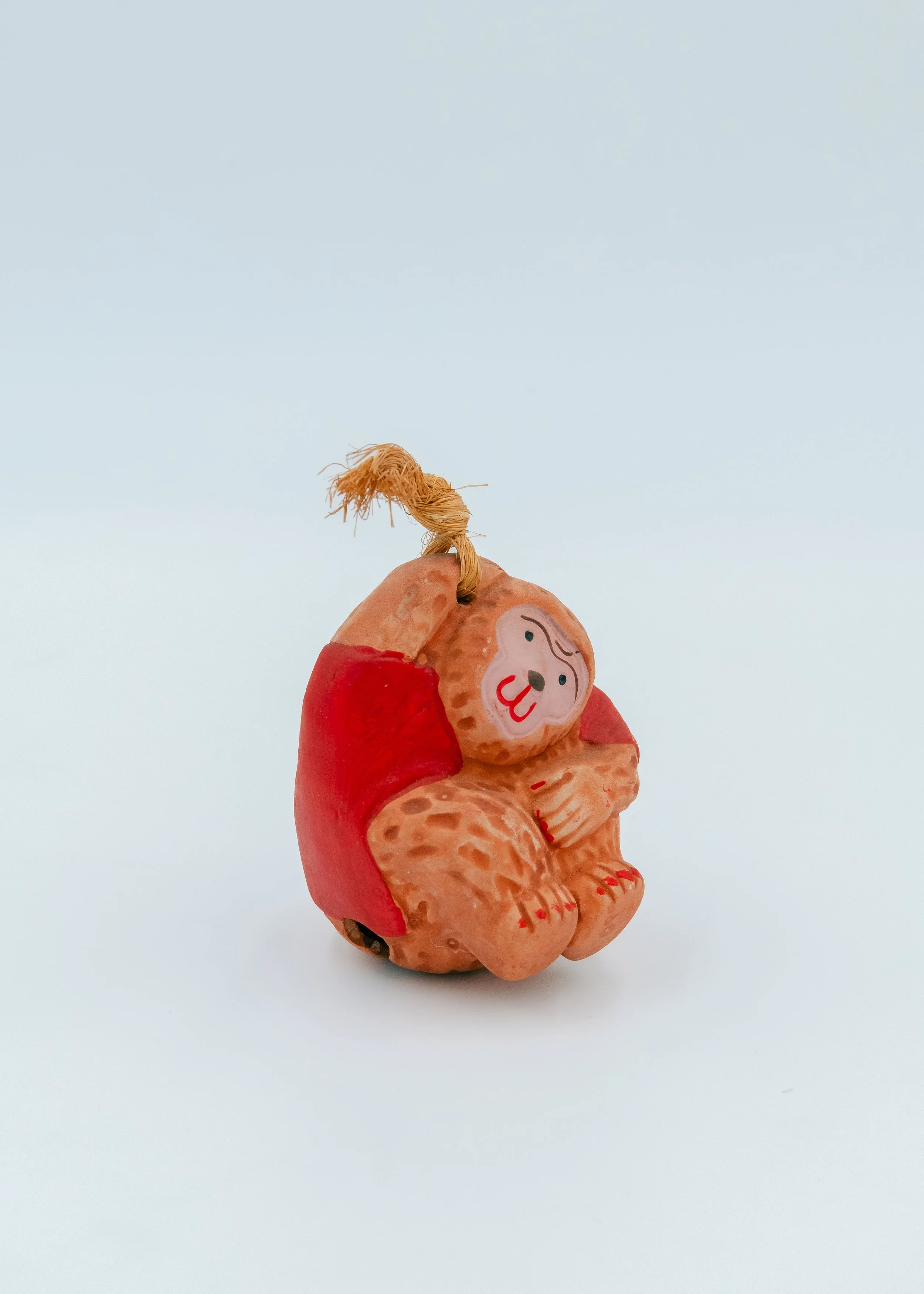 Vintage Japanese clay monkey bell mascot from Saga, wearing a vibrant scarlet hand-painted vest and jute cord, a symbolic amulet for warding off misfortune.