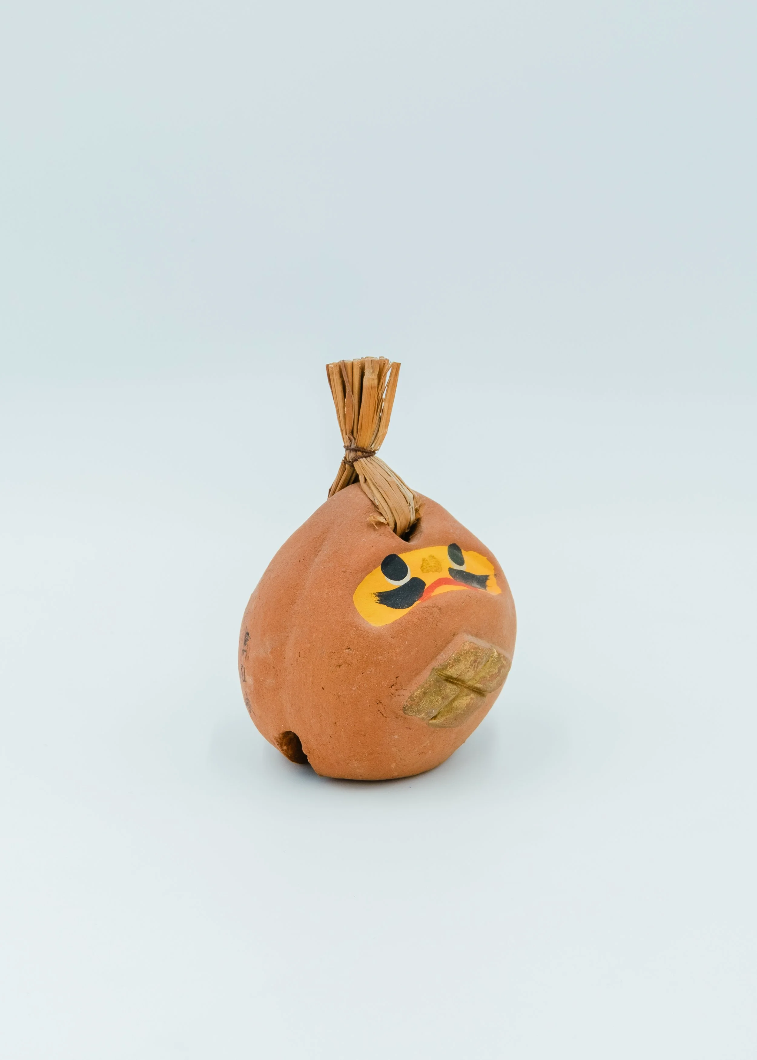 Three-quarter view of UNGLAZED TALISMAN 1970, a rare vintage Japanese earthen bell (Dorei) with hand-painted Daruma face and a traditional straw bundle top.