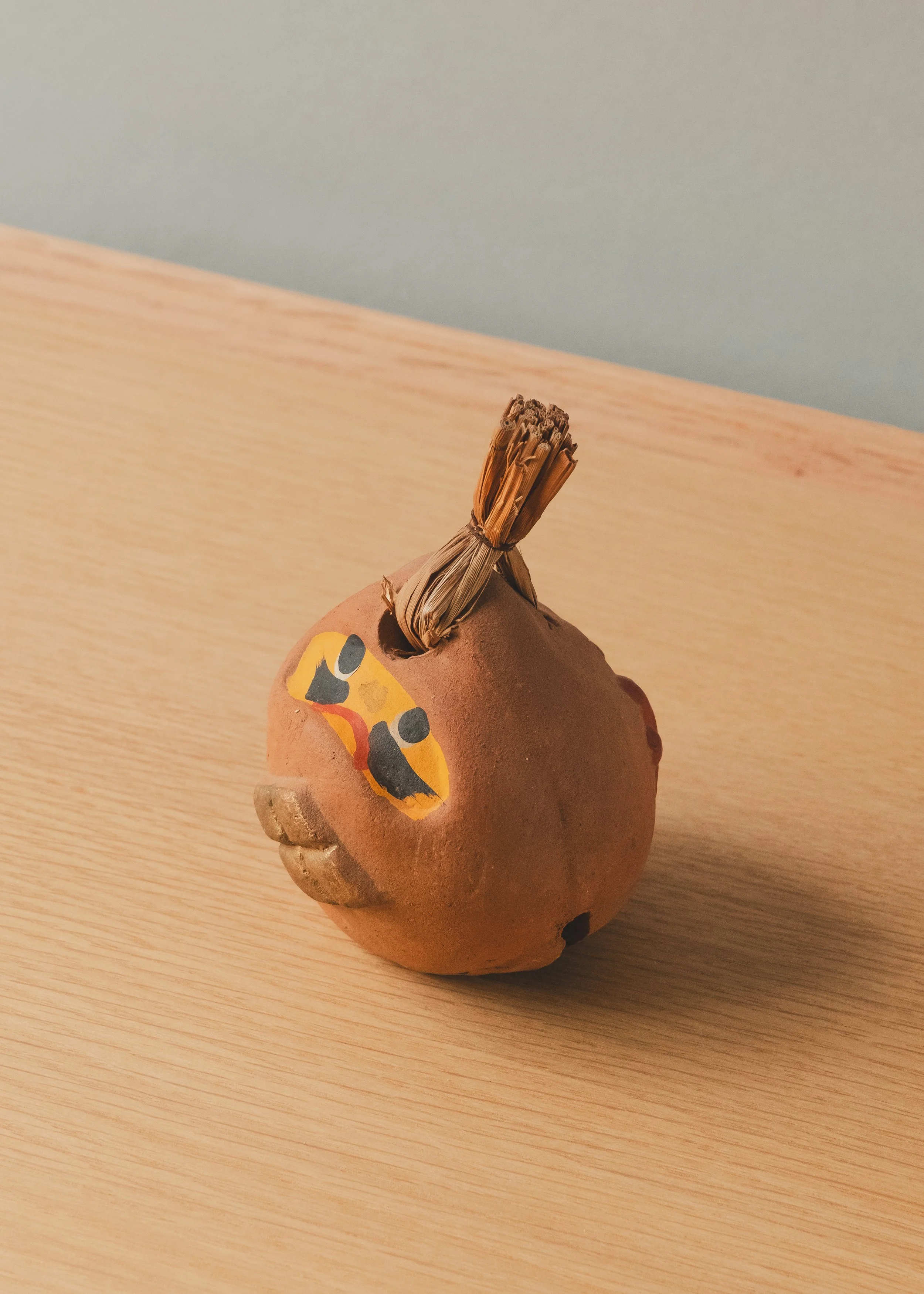 Three-quarter view of UNGLAZED TALISMAN 1970, a rare vintage Japanese earthen bell (Dorei) with hand-painted Daruma face and a traditional straw bundle top.