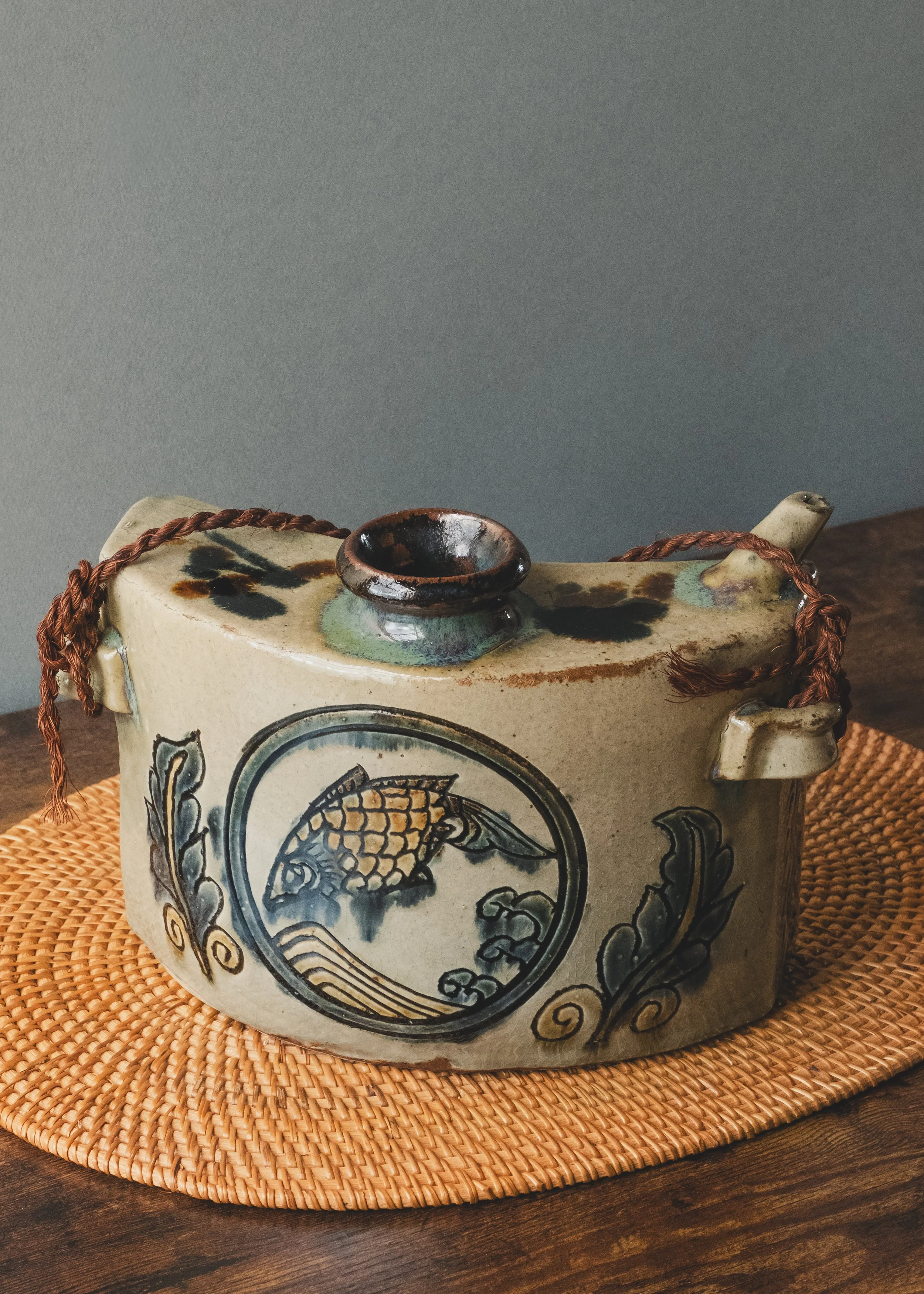 Three-quarter view of a vintage Okinawan Dachibin ceramic bell with fish patterns and a brown ritual cord.