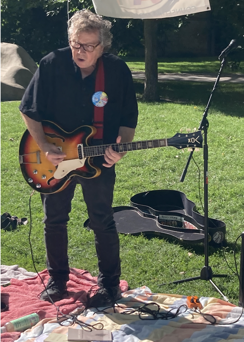 A man with curly gray hair and glasses, wearing a black shirt and black pants, plays an electric guitar outdoors on a red and white blanket, with a black guitar case and a microphone stand nearby.