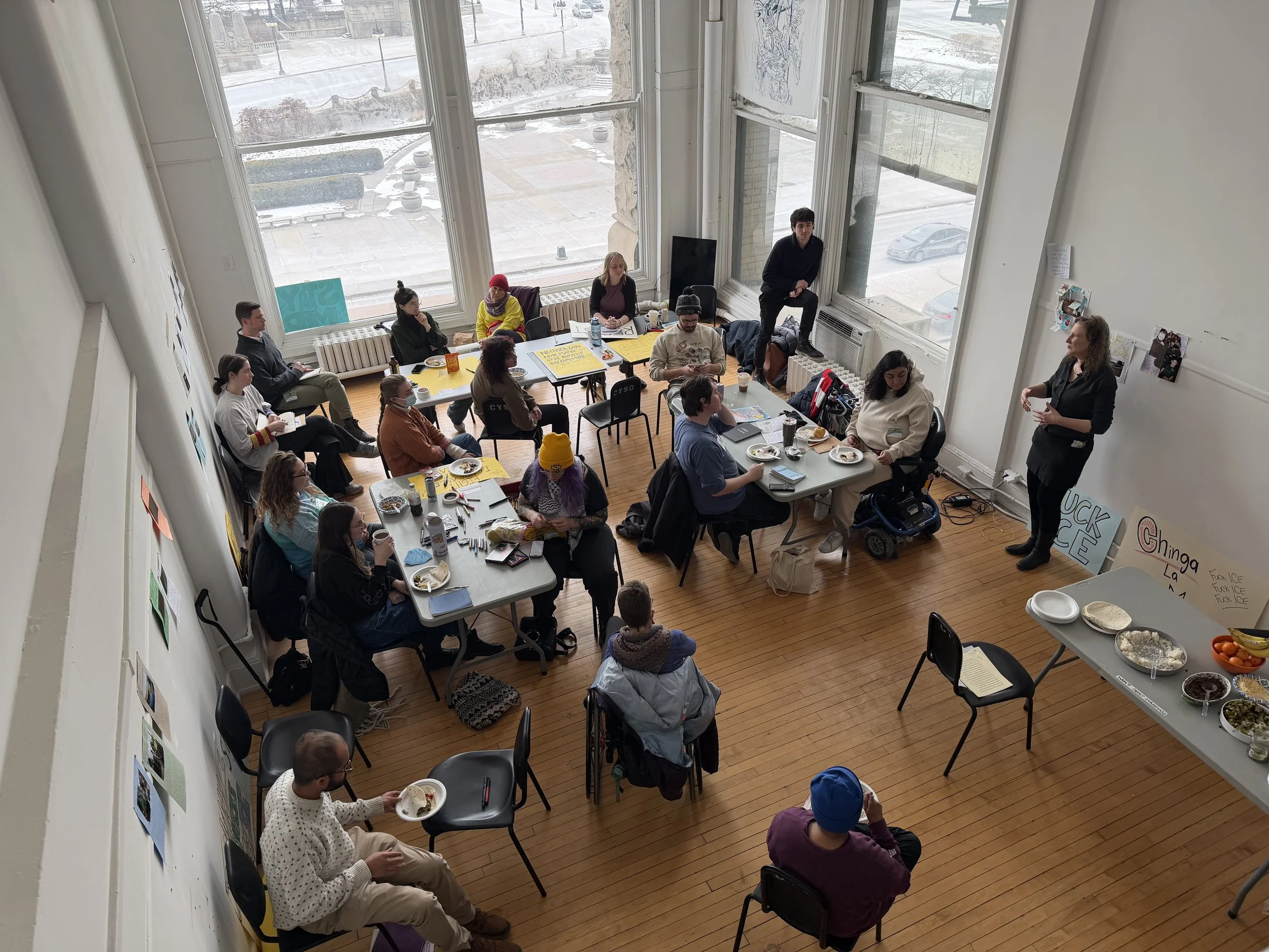 A group of people gathered in a bright room for a meeting or workshop, with tables, chairs, and food on display, and a woman standing and speaking at the front of the room.