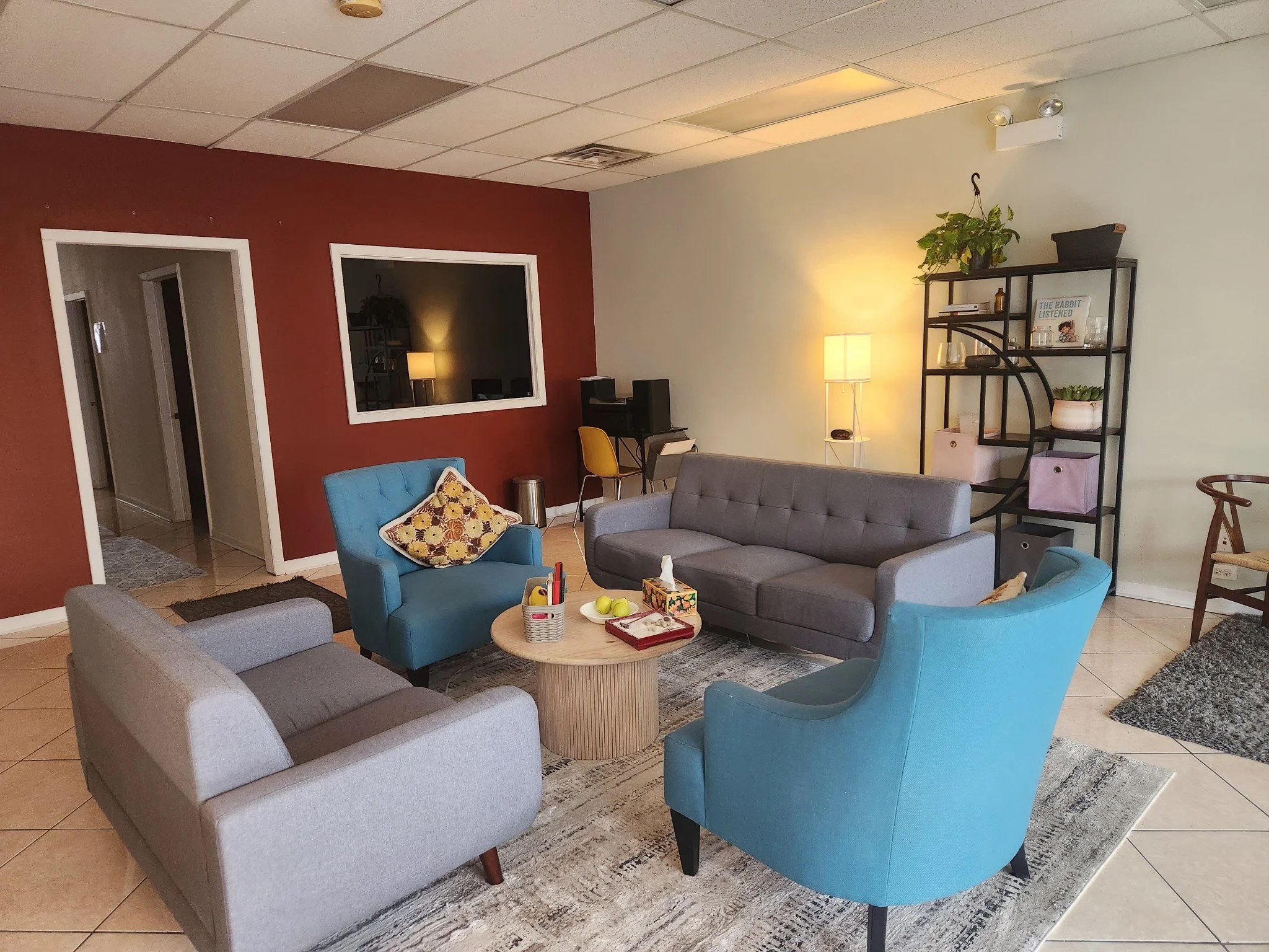 Cozy waiting room with four colorful armchairs arranged around a round wooden coffee table. The room features a maroon accent wall with a window, a black bookshelf with plants and books, and a lamp providing warm lighting. The floor is tiled, and there is a gray and cream area rug under the seating area.