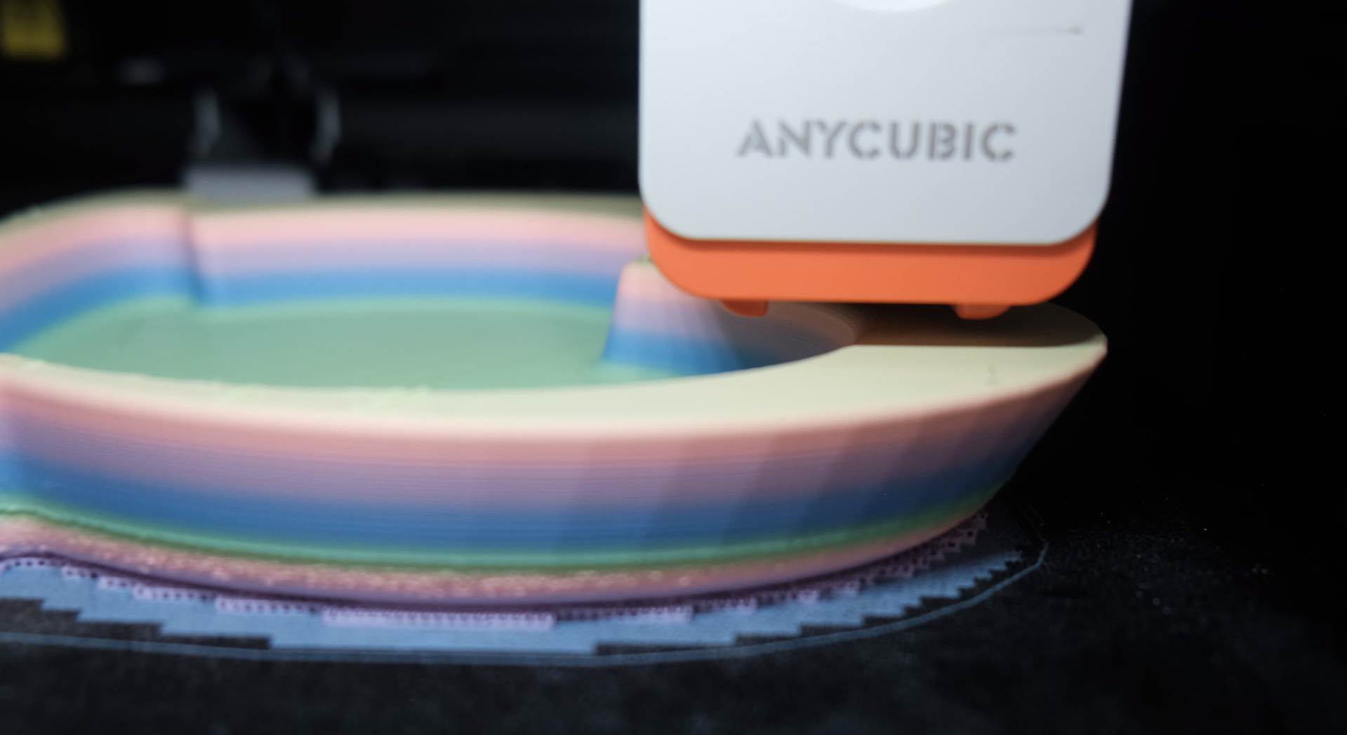 Close-up of a 3D printer extruding colored filament to create a layered rainbow-colored object, with the brand name 'ANYCUBIC' visible on the printer.