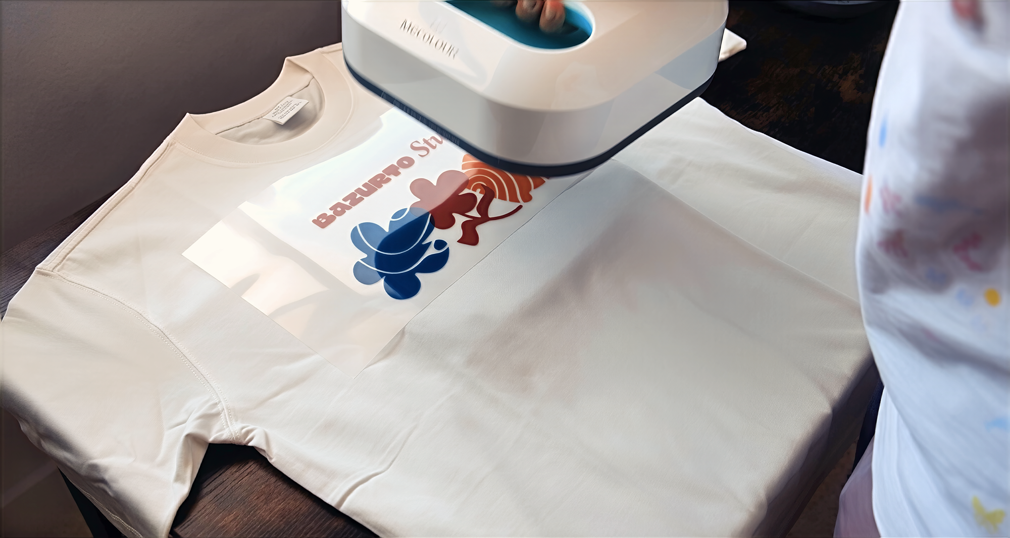 A heat press machine printing a colorful design on a white T-shirt that reads 'BAZURRO St' with a stylized blue and brown flower graphic, on a white tablecloth.