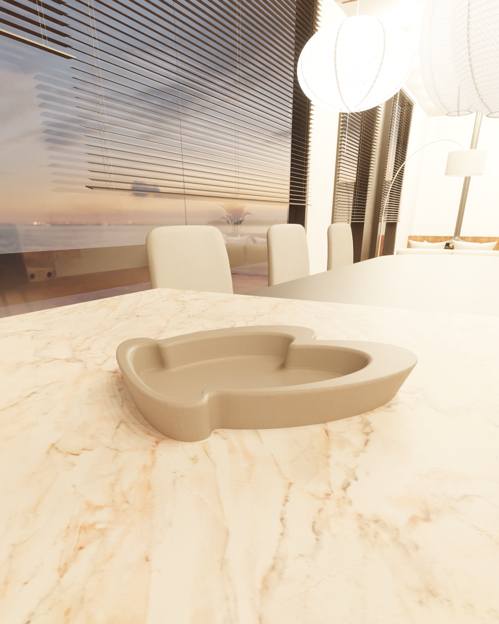 Close-up of a beige marble table with a gray kidney-shaped dish in the foreground. In the background, there are cream-colored chairs, large windows with horizontal blinds, and hanging white spherical pendant lights. The view outside shows a body of water and a sunset sky.