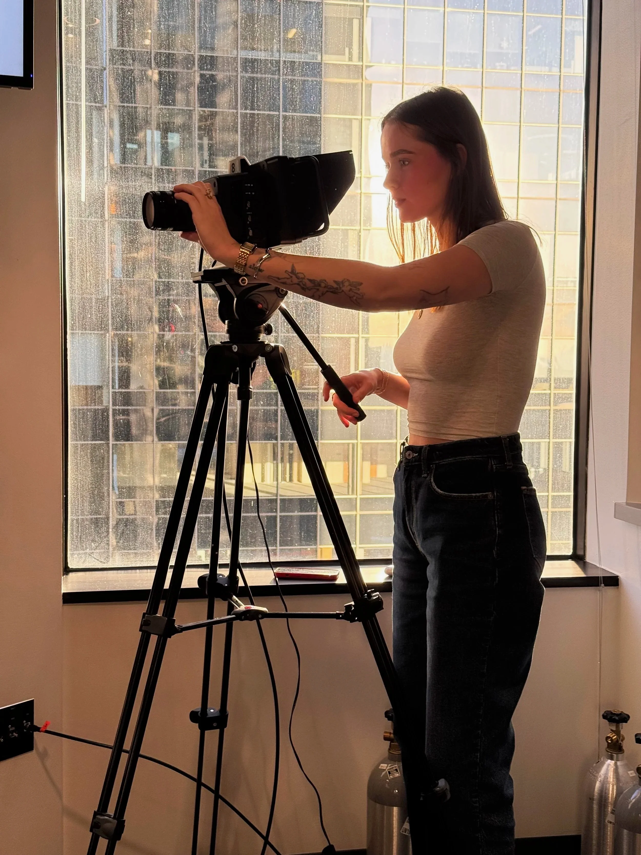 A woman operating a camera on a tripod near a large window in an indoor setting.
